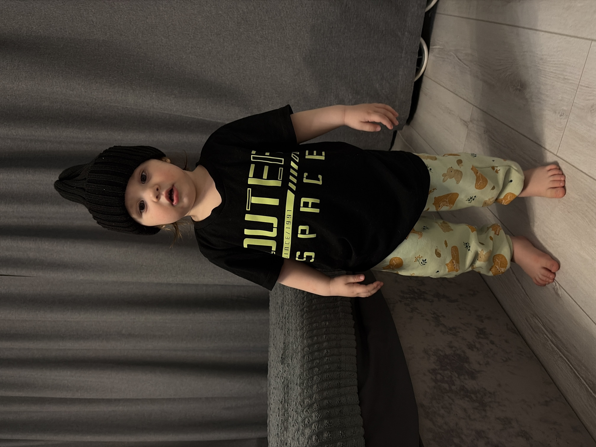 A young child stands indoors, looking directly at the camera with a curious expression and slightly open mouth. The child is dressed in a black knit beanie, a black t-shirt, and light green pants patterned with brown and white animals, likely rabbits and foxes, and small plant motifs. Yellow text on the t-shirt reads "OUTER SPACE" with "SINCE 1991" visible below "OUTER".
The scene is set in an indoor room in Chaikovsky, Russia, possibly a living room or bedroom. The child is standing on a light wooden floor next to a dark grey, textured sofa or couch. A white cable or cord is visible on the floor in the background. The lighting suggests an indoor setting during the day or a well-lit evening. The child's right arm is slightly raised, while the left arm hangs by their side. Their feet are bare.