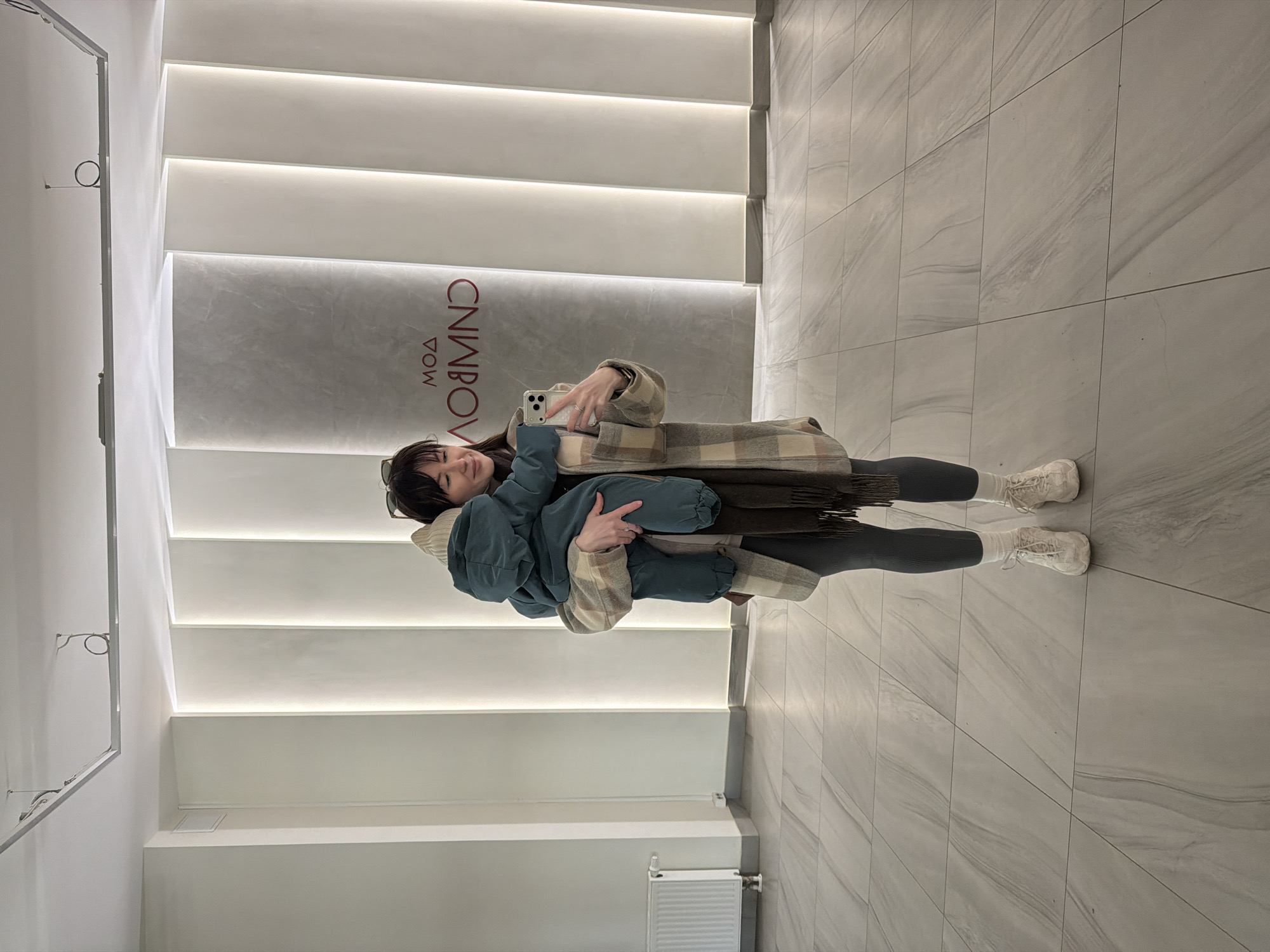 A woman is holding a child and taking a selfie in a modern interior. The woman is wearing black leggings, white socks, and off-white sneakers. She is also wearing a beige and brown plaid coat and holding a brown scarf. The child is wearing a blue hooded snowsuit. The background consists of a white wall with horizontal lighted panels and a gray wall with red text that reads "CNVMODA" in Cyrillic letters. The floor is tiled in a gray marble pattern. The photo was taken in Perm, Russia.