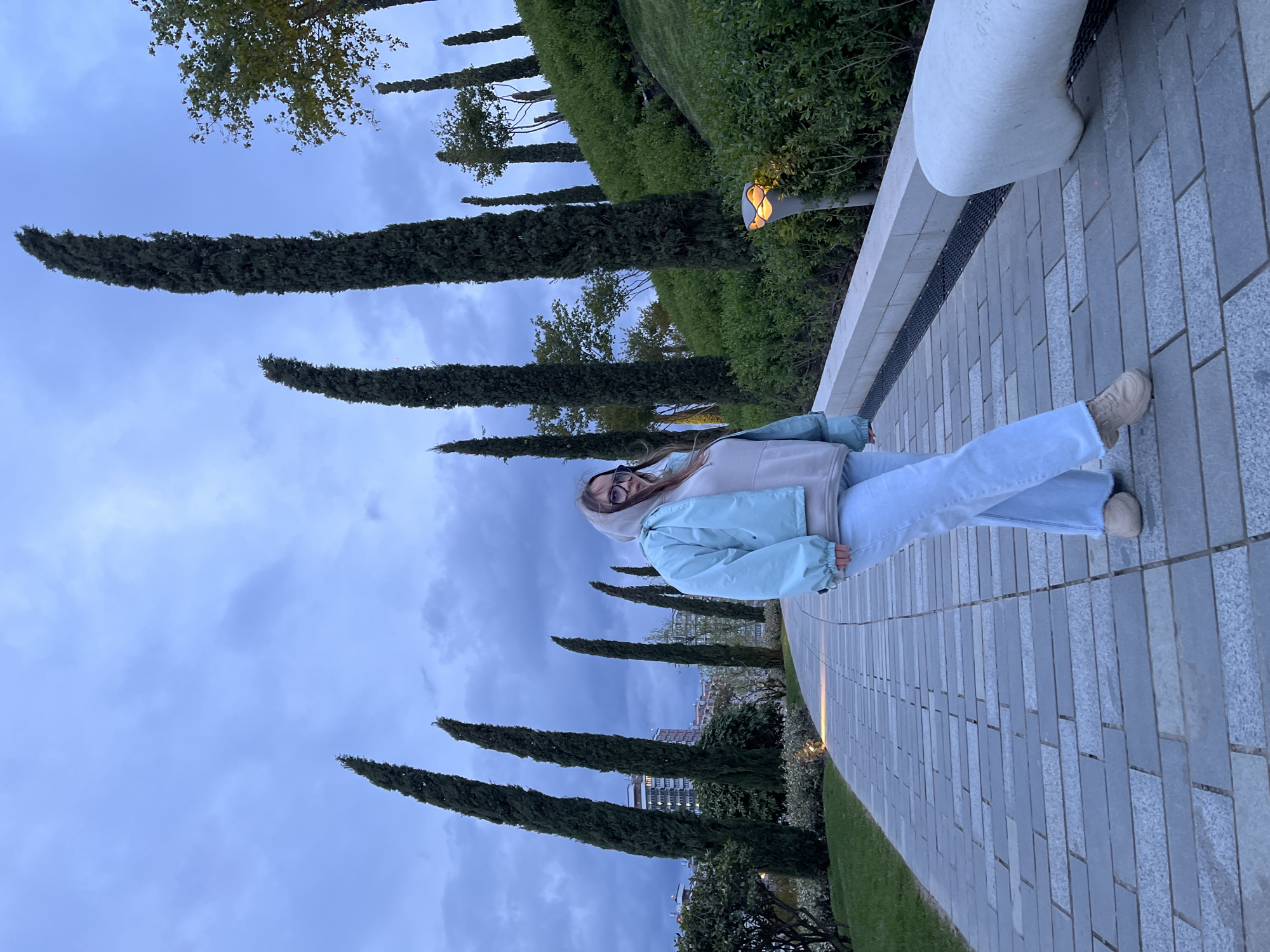 A young woman stands smiling on a paved pathway in an outdoor park in Krasnodar, Russia. She is the primary subject, captured in what appears to be a posed photograph. She wears a light blue puffer jacket with a light-colored hoodie underneath, light blue jeans, and light beige shoes. Glasses are perched on her face.
The setting is a well-maintained urban park or landscaped area. The pathway, made of rectangular light grey paving stones, extends into the background. To her right, a long, light-colored stone bench or low wall runs parallel to the path. Behind this wall, lush green bushes and grass create a soft backdrop. Numerous tall, slender evergreen trees, possibly cypress or juniper, line the path and extend into the distance, with their dark foliage contrasting against the lighter sky. One modern, dome-shaped outdoor lamp is visible among the bushes, emitting a warm glow, suggesting the time is late afternoon or early evening. Distant buildings can be partially seen through the trees.
The sky is overcast with soft, diffused clouds, indicating cloudy weather. The overall light is dim but even. The woman's smile conveys a happy or content emotion. There is no visible text in the image.