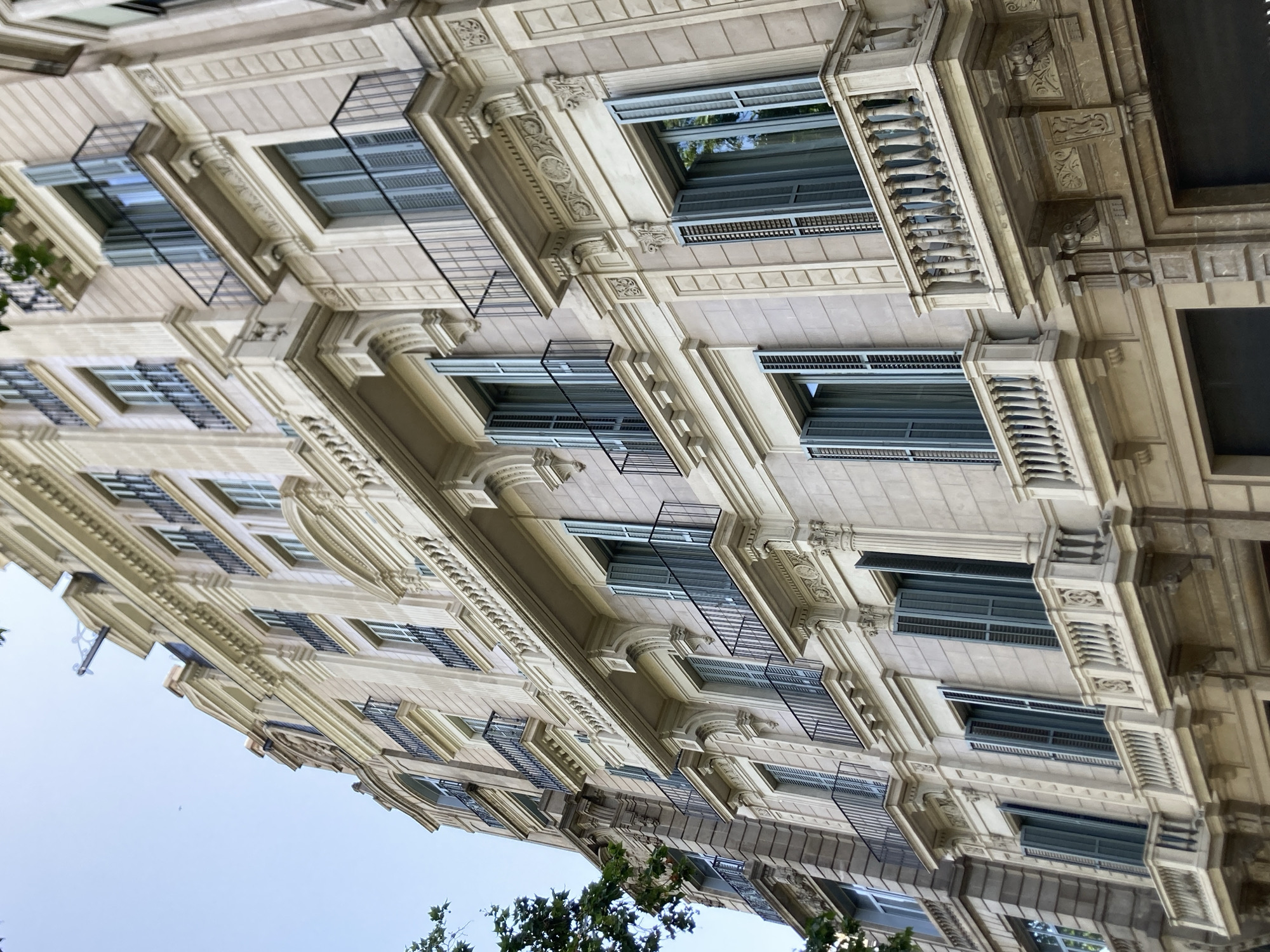 This is a low-angle, close-up, exterior shot of a light-colored building with multiple stories. The building's facade is ornate, featuring detailed carvings, pilasters, and balconies with decorative railings. Windows with grey shutters are visible on each story, set within recessed frames. The overall architectural style suggests a classic European aesthetic, possibly from the late 19th or early 20th century. The image is oriented vertically, giving the impression of looking up at the building from a street level. Green foliage from trees is visible at the bottom of the frame, suggesting the building is located in an area with trees. The sky is a pale, overcast grey, indicating it is likely daytime, possibly an overcast day. The building appears to be residential or a hotel, given the windows and balconies. There is no visible text in the image. The scene is peaceful and evokes a sense of urban architectural beauty. The building's detailed craftsmanship is a prominent feature.