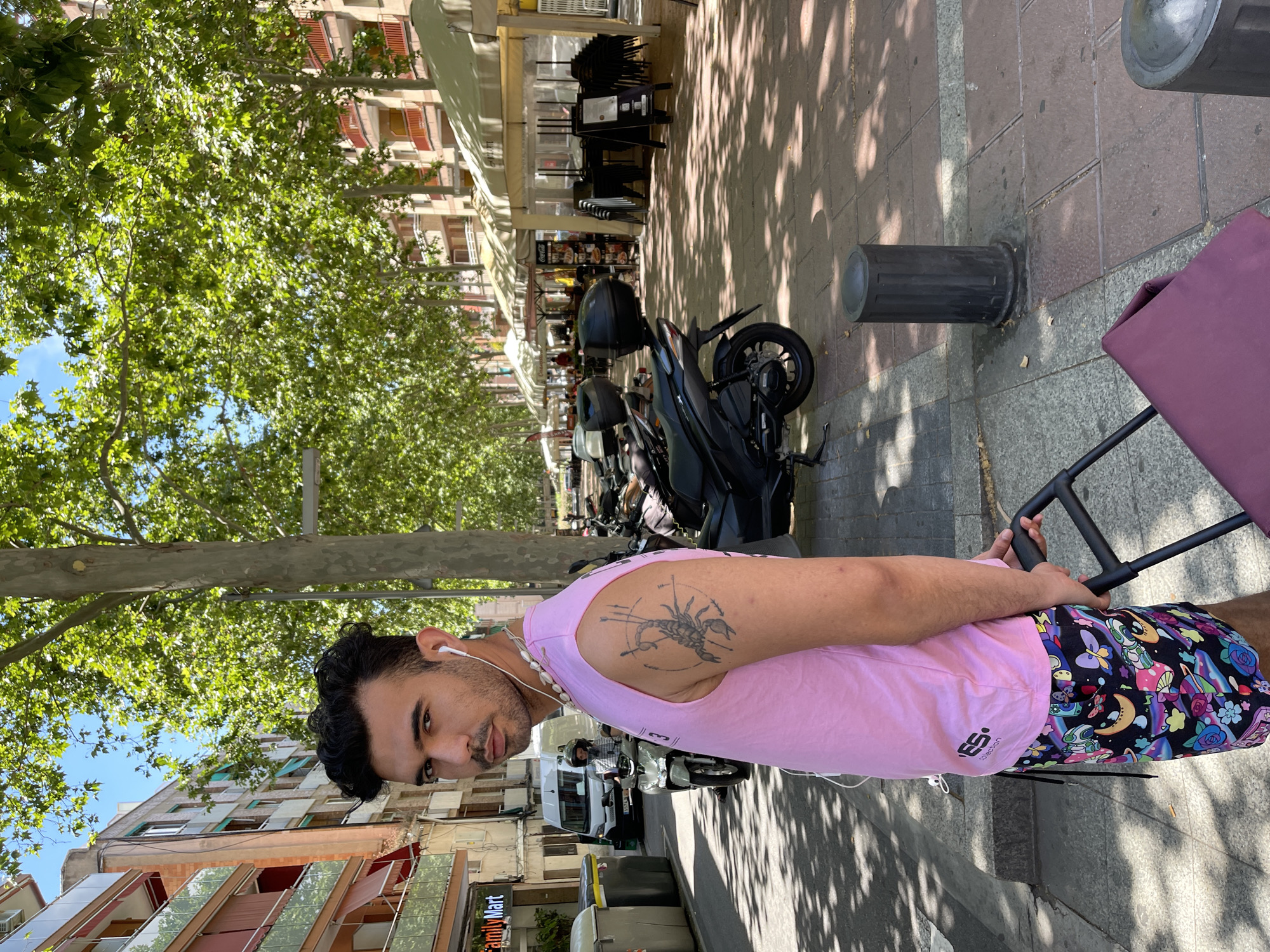 A young man with dark hair and a beard stands on a sidewalk in Santa Coloma de Gramenet, Spain, pulling a shopping cart with a purple bag. He wears a pink sleeveless shirt with a scorpion tattoo on his left arm, and colorful shorts featuring cartoonish designs. He has white earbuds in his ears and a necklace made of shells. Behind him, a street lined with trees and apartment buildings is visible. Several parked motorcycles and a white van are also present, along with outdoor seating areas of a restaurant. The scene appears to be a sunny day, with shadows cast by the trees on the pavement.
