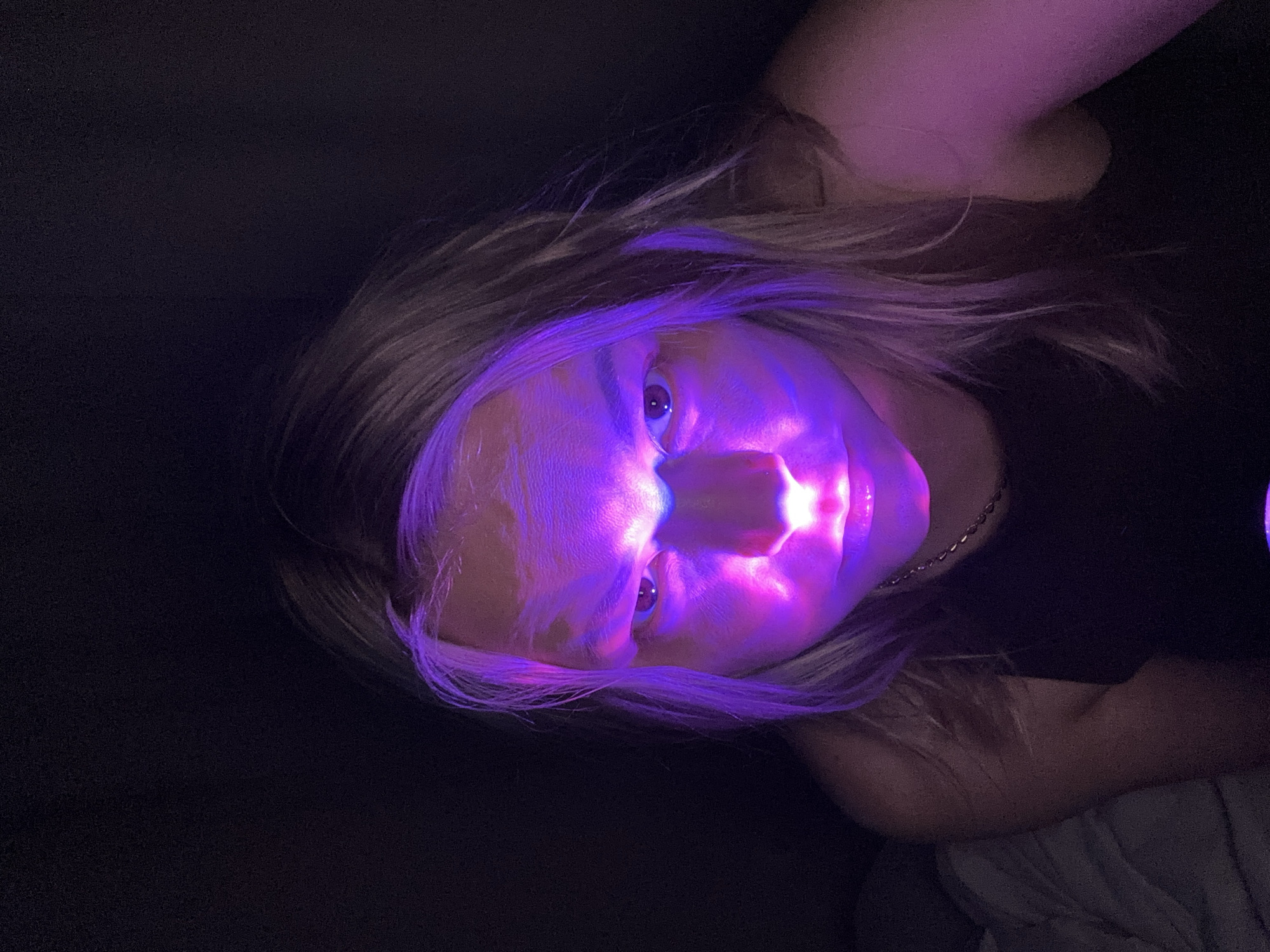 A portrait of a person with fair skin and light hair is illuminated by a strong purple light, creating stark contrasts between the lit face and the dark background. The light source appears to be close to the face, emphasizing features like the nose and mouth with intense brightness. The background is predominantly dark, with hints of the person's clothing. There is no indication that this image was taken in Izhevsk, Russia, or any other specific location.
