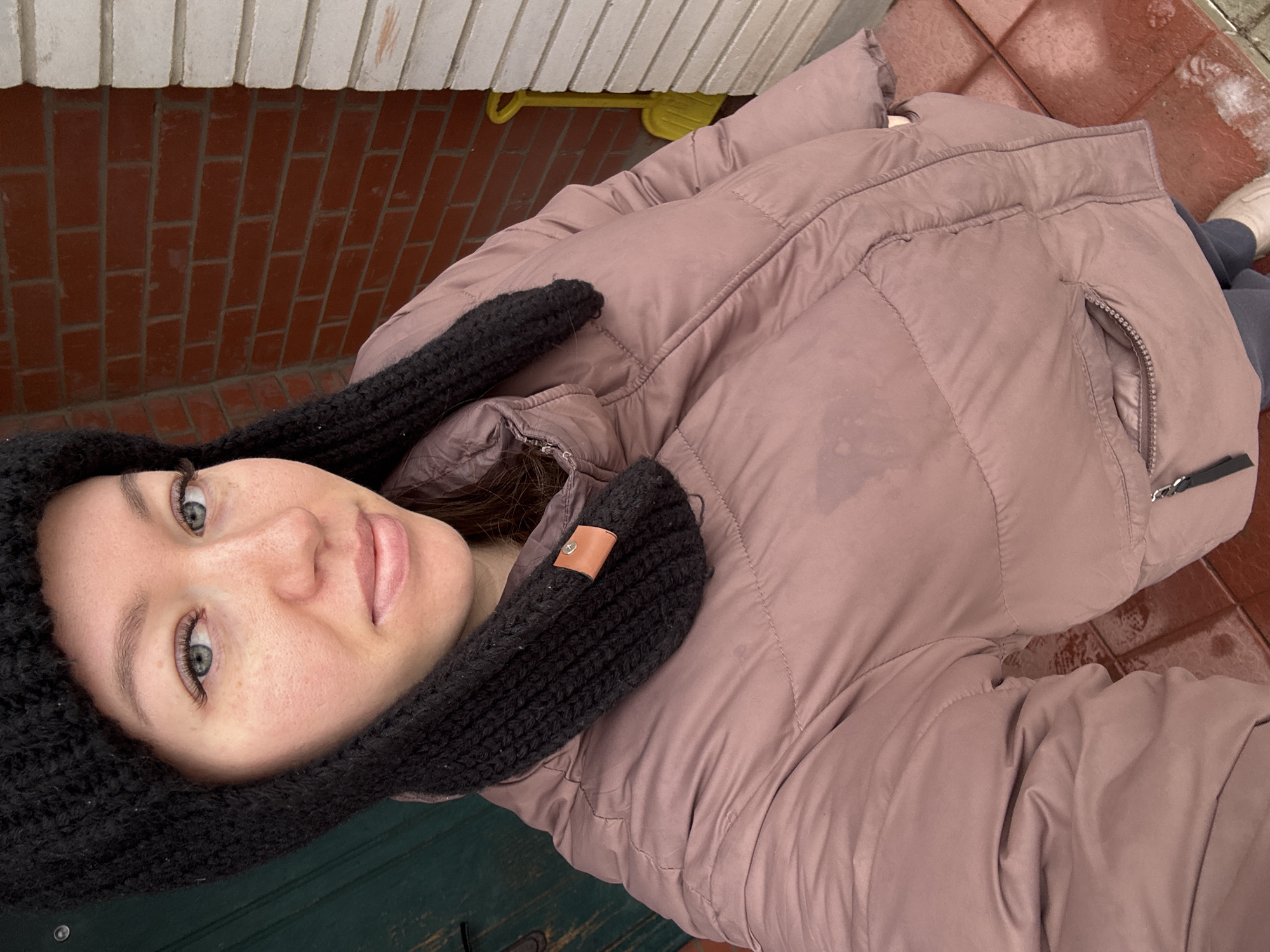 A woman with fair skin and blue eyes, wearing a black knit hat and a mauve-colored puffer jacket, is looking directly at the camera. The scene appears to be outside, near a building with red brick and an adjacent area with red tiles. A partial view of a yellow child's shovel is visible in the upper left corner. The setting is in Ферма, Russia.