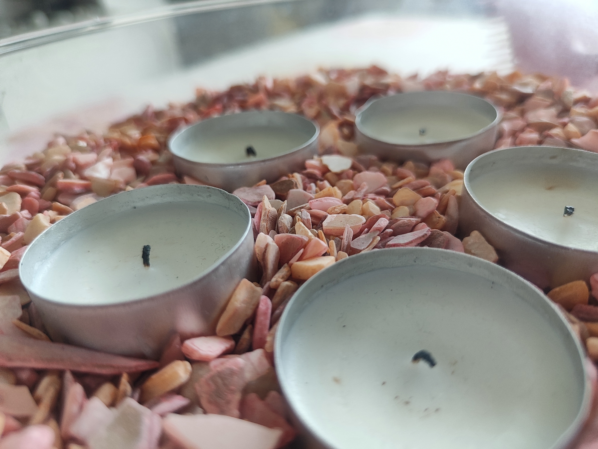 A close-up, eye-level shot shows four unlit tea candles with black wicks, nestled in a bed of small, irregularly shaped, multi-colored pebbles and fragments. The pebbles are predominantly shades of pink, beige, and light brown. The candles are housed in silver-colored metal holders. The background is blurred and appears to be a light-colored, possibly reflective surface, with some vertical lines visible at the top. There is no discernible indication of the location in Voronezh, Russia.