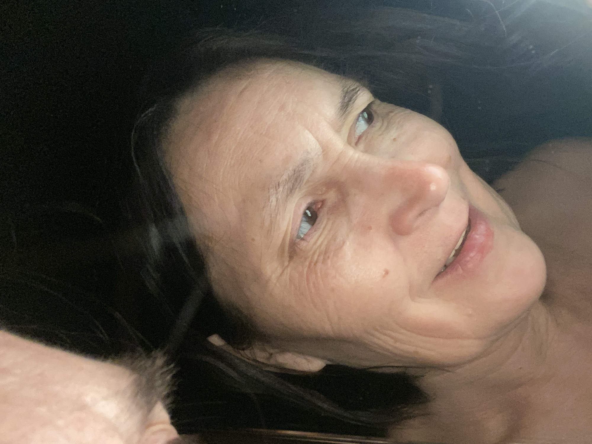 An older woman with dark hair is looking upwards with a slight smile. Part of a second person's head is visible in the lower left corner of the frame. The environment is dimly lit. The scene does not appear to be located in Novokuznetsk, Russia, as there is no discernible context to place the image there.
