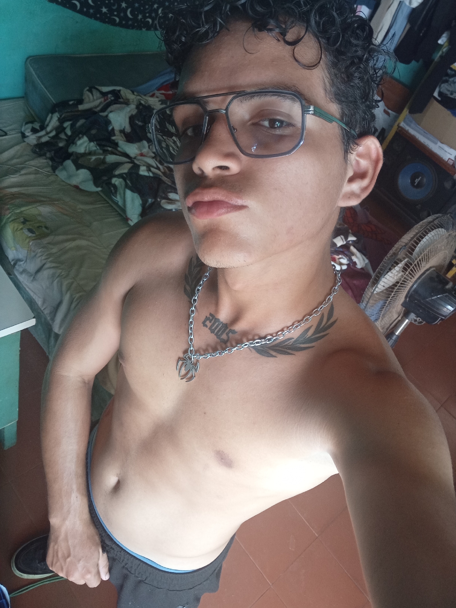 A young, shirtless man with curly dark hair and glasses stands in what appears to be a bedroom in Altagracia, Venezuela. He is looking at the camera, with his lips pursed. He is wearing a silver chain with a spider pendant and has tattoos on his chest and shoulder. In the background, a bed with patterned sheets is visible, along with a fan and some clothing. The floor is tiled.