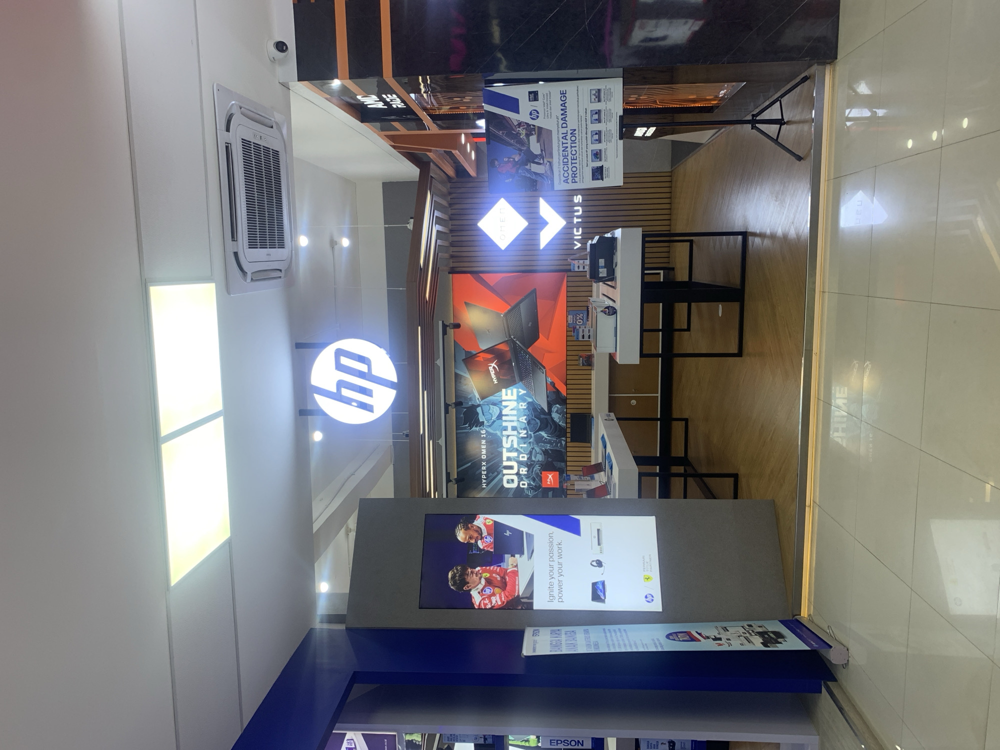 The image shows an HP store in Palembang, Indonesia, likely located within a shopping mall given the clean, modern interior and tiled flooring. The store features a prominent HP logo illuminated from above. On the left, a large digital display showcases an advertisement with the tagline "Ignite your passion, power your work." The advertisement features two men in racing attire looking at a laptop, suggesting a partnership with a racing team. Below this, there are images of a laptop, headphones, and a desktop computer. Further back, a large screen displays HP Omen laptops with the slogan "OUTSHINE ORDINARY." Several HP laptops are displayed on white tables. To the right, there is a sign for "ACCIDENTAL DAMAGE PROTECTION" and other informational posters. An air conditioning unit is visible on the ceiling, and a security camera is mounted high on the wall. The overall environment is well-lit and organized, typical of a retail electronics store.
