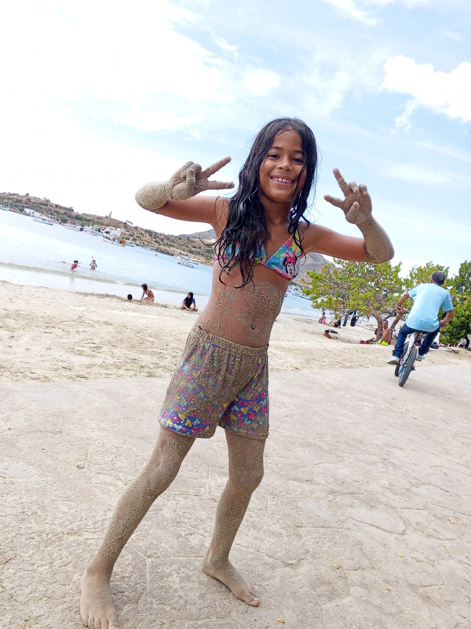 A young girl covered in sand stands on a beach on the island of Margarita, Venezuela, giving a peace sign with both hands. The girl is wearing a multicolored bikini top and shorts. In the background, there is a beach with people swimming and sunbathing, as well as a man riding a bicycle. The sky is blue with some clouds.