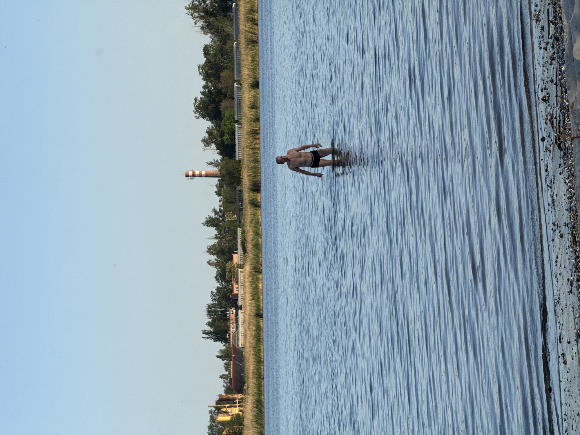A man in black swim trunks stands in shallow water. To the left of the water, there is a shoreline with sparse vegetation, buildings, and a red and white chimney. A pale blue sky is visible above the landscape on the left. The shoreline to the right is covered in pebbles. The location is likely near Rostov-on-Don, Russia.