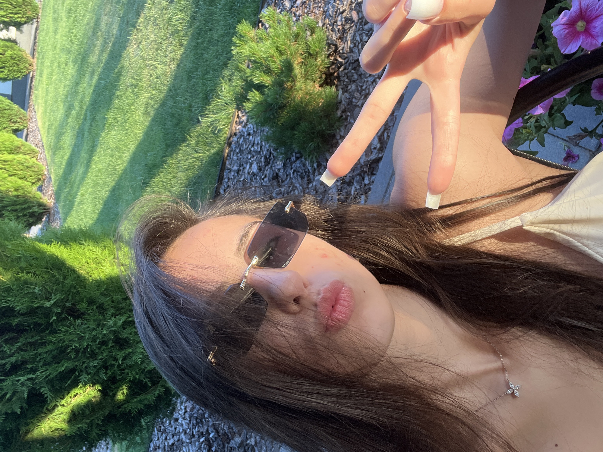 In Novooleksandrivka, Ukraine, a young woman with long, dark hair and sunglasses is taking a selfie outdoors. She is wearing a light-colored top and a delicate silver necklace. Her lips are pursed, and she is making a peace sign with her right hand, which has long, white-painted nails. The background features a manicured lawn, trimmed evergreen bushes, and shadows cast by sunlight, suggesting it is daytime.