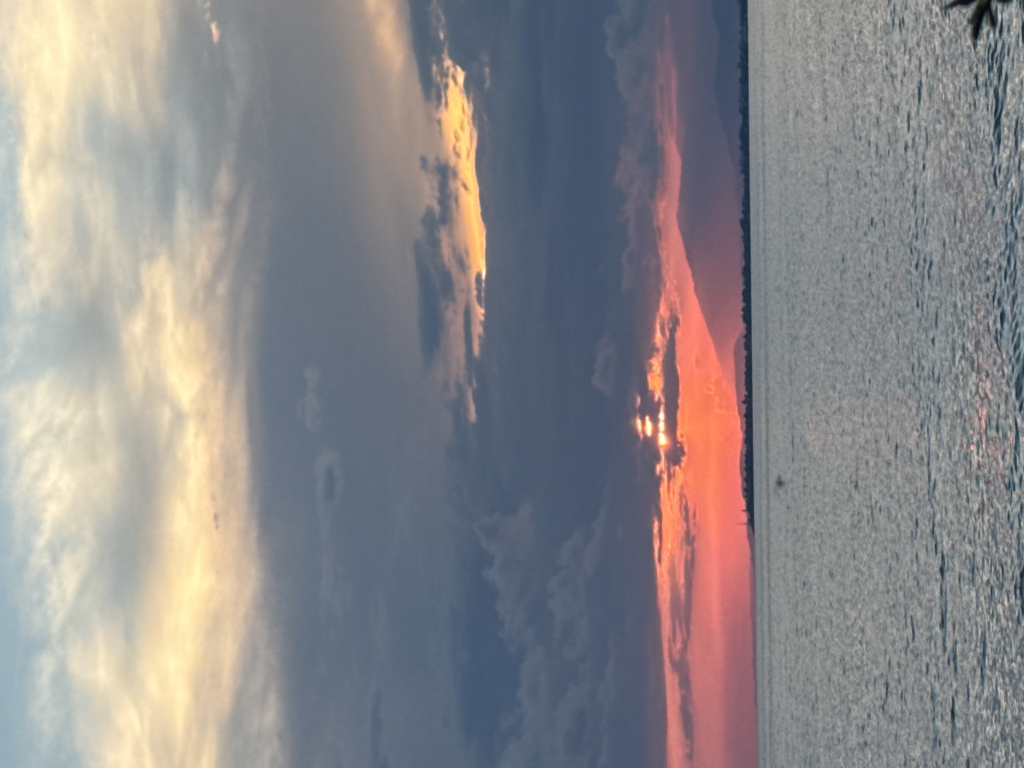 A sunset over the sea is depicted. The sky is filled with dramatic clouds, ranging from light yellow and orange hues near the horizon to dark gray and blue at the top. The sun is partially obscured by clouds, casting warm light through gaps. In the distance, a silhouetted mountain range borders the horizon, with a faint outline of a city or town visible on the left. The sea in the foreground is calm, reflecting the colors of the sky. The image appears to have been taken from a low vantage point, likely at sea level. There are no people or specific identifiable objects in the foreground. The scene evokes a sense of natural beauty and tranquility.