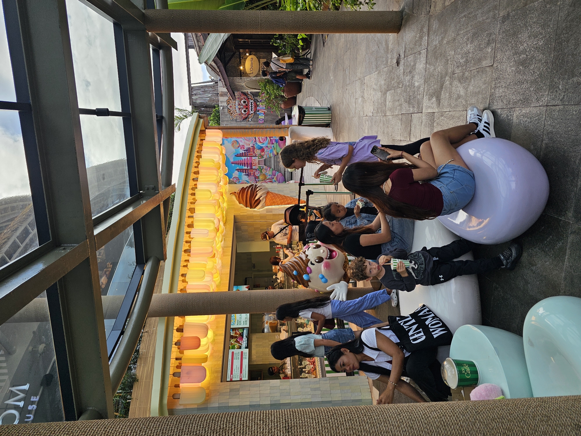 The image depicts an outdoor seating area in Denpasar, Indonesia, likely at a commercial establishment such as a cafe or ice cream parlor. A row of illuminated pastel-colored lamps hangs above a storefront with a bright yellow facade. A large inflatable ice cream cone character stands near the entrance. Several people are seated on oversized, bulbous white and light purple seats in the foreground and midground.
In the foreground, a woman in a black top and white shirt is seated on a white pouf, with a black tote bag that reads "GENTLE WOMAN" on her lap. To her right, a young boy with curly hair is seated on a white pouf, holding a green and white striped cup with a straw and looking towards the camera. Next to him, a young woman in a maroon top and denim shorts is seated on a light purple pouf, looking at her phone.
In the midground, to the left of the ice cream character, two young women are standing, one in a light blue top and denim skirt, and the other in a white t-shirt and blue pants. To the right of the ice cream character, a young woman in a purple top and white sneakers is interacting with the woman in the maroon top. Further in the background, several other individuals are visible, including a couple standing near an establishment labeled "L'Osteria" and other patrons seated. The environment is paved with stone tiles, and there are large, textured pillars supporting a modern overhead structure with glass panels. Lush greenery is visible in the background. The lighting suggests it is either late afternoon or early evening.