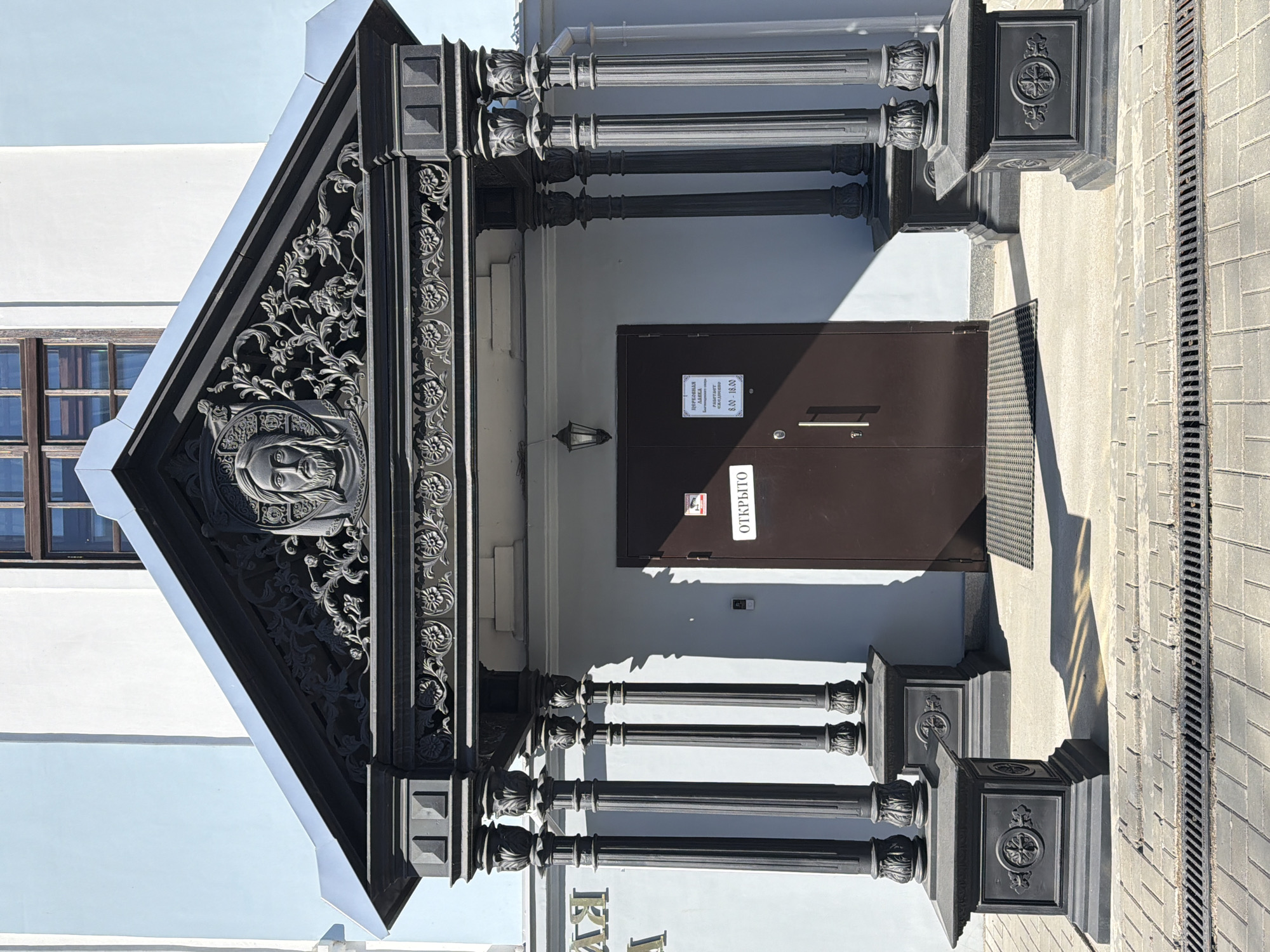 The image shows the entrance to a building, likely a church, located in Votkinsk, Russia. The entrance is ornate, featuring a triangular pediment with decorative carvings, including a religious icon depicting a face. The entrance is framed by columns, the capitals and bases of which are adorned with floral designs and crosses. A dark brown double door is recessed within the entrance, with signage in Russian indicating opening hours and stating "Open." The building's exterior is painted in light blue and white. The sidewalk in front of the building is paved with gray rectangular stones and includes a drainage grate.