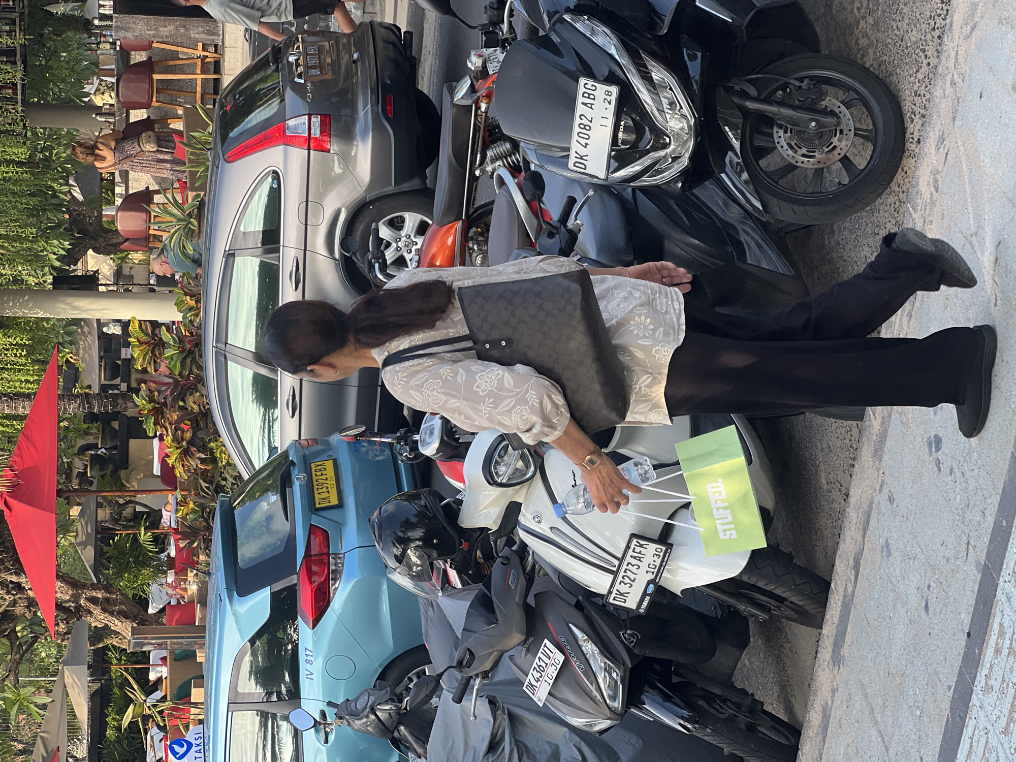 In Kuta, Indonesia, a woman in a white patterned blouse and black pants walks past several parked motorbikes. She carries a dark patterned handbag over her right shoulder and holds a water bottle and a bright green shopping bag in her left hand. The shopping bag has "STUFFED." printed in white letters. The motorbikes are parked on a sandy area next to a paved walkway. Behind the motorbikes, a light blue car and a dark gray car are visible. In the background, red umbrellas shade outdoor seating areas with people relaxing. The environment suggests a commercial or tourist area.