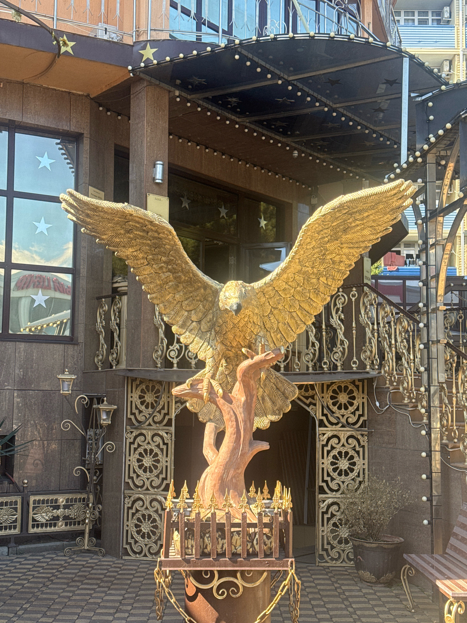A large, golden eagle sculpture is positioned centrally in front of a building in Anapa, Russia. The eagle is depicted with its wings spread wide and is perched on a brown, tree-like base. The building itself has a dark brown facade and features large windows with white star decorations. Above the entrance, a dark canopy is adorned with strings of small, white lights and decorative stars. Ornate gold railings and decorative metalwork are visible along the stairs and around the building's entrance. The ground is paved with textured tiles. A dried potted plant sits to the right of the entrance, next to a wooden bench with gold accents.