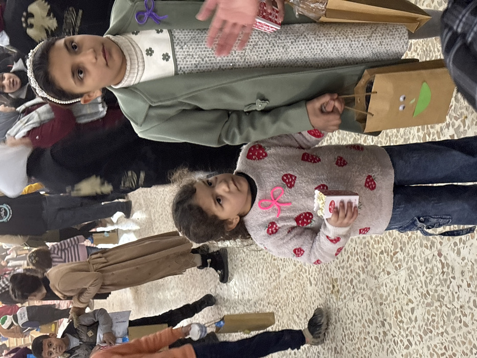 The image depicts an indoor gathering in Kafr Batna, Syria, featuring a crowd of individuals, with two girls prominent in the foreground. The flooring is a light-colored terrazzo or tiled surface.
In the right foreground, an approximately 10-14-year-old girl with dark hair and a pearl-studded headband looks slightly to her right. She wears a light olive-green long coat over a white turtleneck sweater featuring a patterned design with small grey motifs and metallic embellishments. A purple ribbon bow is pinned to her upper left chest. Her left hand holds a brown paper bag adorned with googly eyes and a green paper mouth. A partial view of another hand, belonging to an unseen person to her right, holds a small red and white box and a second plain brown paper bag.
To the left and slightly in front of the older girl, an approximately 4-6-year-old girl with dark, curly hair looks upwards and to her left with a serious expression. She wears a light beige fluffy long-sleeved sweater embroidered with multiple red strawberries and a prominent pink ribbon bow appliqué on the chest. She is also wearing dark blue cargo-style jeans. Her right hand holds a small white and red box filled with popcorn.
In the midground and background, numerous blurred figures are visible. On the left, a person, possibly a boy, holds a small white cup and a brown paper bag. Further back, another person in a long tan coat and dark boots is partially visible. Several adults and children are present, with some adults wearing head coverings. A green and white logo displaying Arabic script (reading "رحمة") is visible on the back of a dark garment worn by an adult in the background.