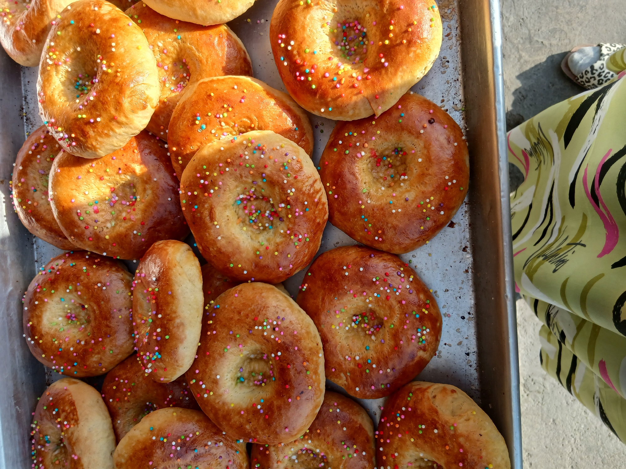 In Khankiz, Uzbekistan, a tray filled with freshly baked, circular bread decorated with colorful sprinkles sits on a weathered metal surface. The bread, golden brown with a slightly uneven texture, features a central opening and is generously topped with a vibrant mix of red, blue, yellow, green, and white sprinkles. The tray, showing signs of age and use, is partially visible as it rests on a rough, textured ground. In the foreground, a person's legs are visible, wearing a patterned skirt in shades of green, black, and pink, and a sandal with a leopard print. The lighting suggests an outdoor setting, possibly a market or a food stall, with shadows cast on the ground.