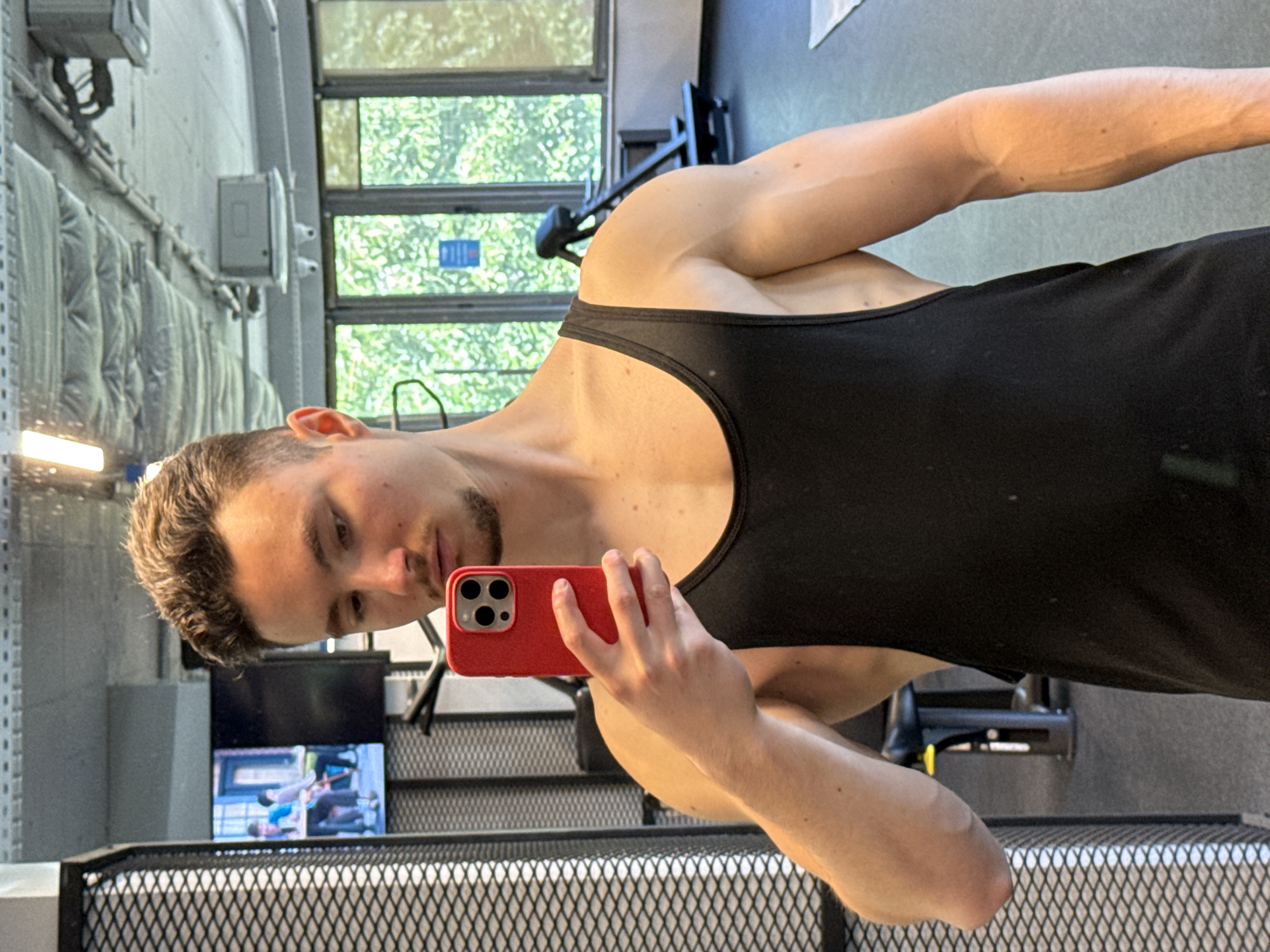 A Caucasian male with short brown hair and a light beard is taking a selfie in a gym. He is wearing a black tank top. The gym has windows overlooking greenery, and exercise equipment is visible in the background. A television displays people exercising. The location is likely in Sofia, Bulgaria, but this cannot be confirmed with certainty from the image alone.