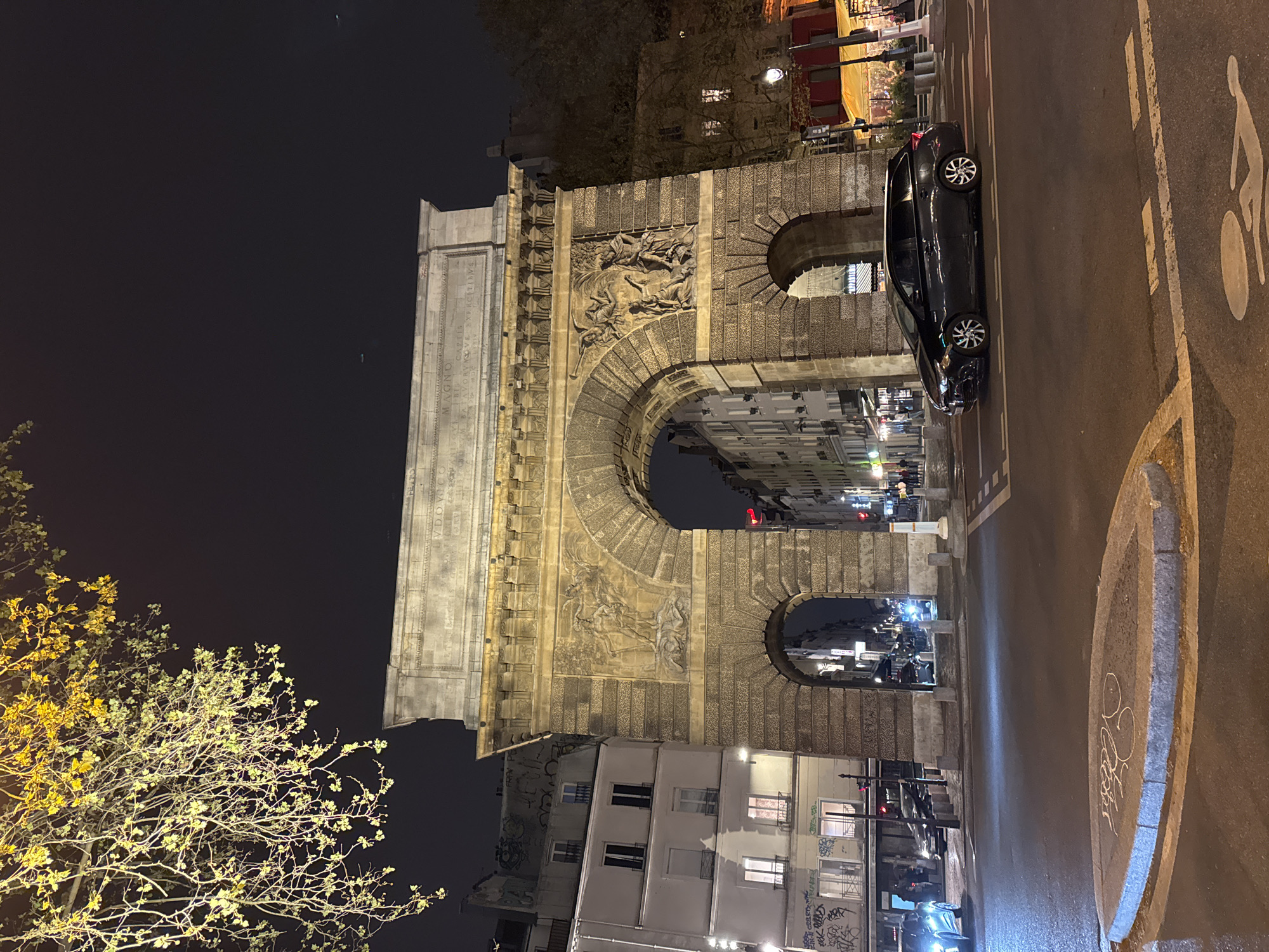 The image captures a nighttime urban scene centered on the Porte Saint-Denis, a triumphal arch located at the intersection of Rue Saint-Denis and Rue du Faubourg Saint-Denis in Paris, France. The large stone arch, constructed from ashlar blocks, features a central large archway and two smaller pedestrian passages. Its facade is adorned with intricate bas-relief sculptures depicting classical figures and military scenes, with a prominent inscription panel visible at the top.
The sky is dark, indicating night, with a tree branch featuring illuminated yellow-green leaves hanging into the upper left of the frame. To the left of the arch are multi-story residential or commercial buildings with white facades, dark windows, and some visible graffiti on lower walls. To the right, buildings line the street, including one with a red awning and warm interior lighting, possibly a restaurant or shop.
In the foreground, an asphalt road displays white lane markings, including a painted bicycle symbol indicating a bike lane. A small, elongated concrete median with faint white chalk graffiti is positioned centrally in the foreground. A dark-colored station wagon or hatchback, likely a Toyota, is positioned on the road in the mid-ground, facing left, with its wheels turned slightly. Distant vehicles with illuminated headlights are visible on the left side of the road and through the central archway. Several indistinct pedestrian figures can be seen walking on the sidewalks on both sides of the street and through the arch. Street lamps emit a warm yellow light, illuminating parts of the arch, buildings, and the wet-looking road surface, with traffic lights showing red in the distance.