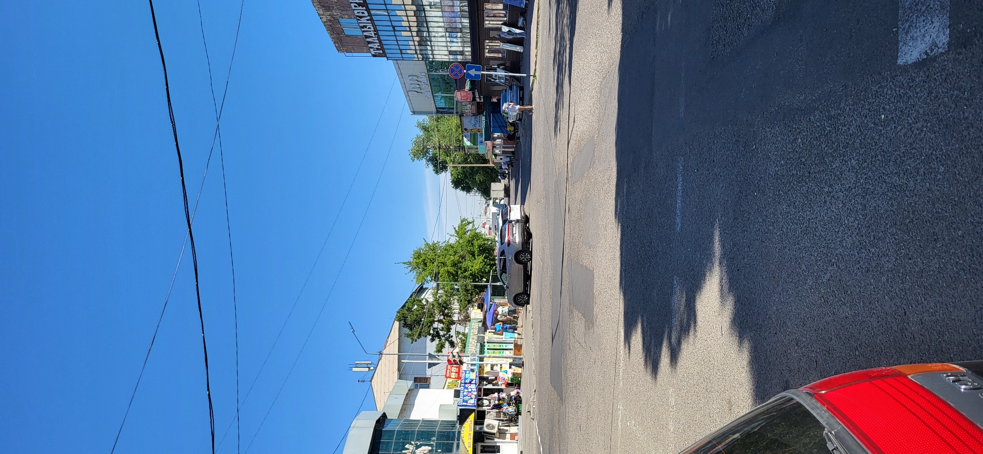 The image captures a street scene in Taldykorgan, Kazakhstan, likely on a clear, sunny day. The perspective is from a street level, looking across an intersection. In the foreground, the asphalt road surface is visible, with shadows of unseen objects casting across it.
To the left, a modern building with glass facades is partially visible, adorned with signs that appear to be in Russian or Kazakh, with text such as "ВСТ. ДЛЯ ШТИН" visible. Adjacent to this are several smaller market stalls or shops, some with awnings and displays of goods. People are seen browsing or working at these stalls.
In the center of the frame, a silver SUV is stopped at the intersection, which is marked with pedestrian crossings. A traffic light with a red signal is visible above the intersection. Further down the street, more buildings and market structures line the road.
To the right, a larger building with a facade that has a metallic sheen and Arabic script-like lettering, likely the name of a business, is prominent. The signage above this building includes the word "ТААДЫКОРП". Below this, there is another section of shops, some with blue awnings. A street sign with a blue arrow indicating a one-way direction is also present.
On the right side of the street, several individuals are walking and standing, and a few more market stalls are visible. Trees with green foliage are scattered throughout the scene, contributing to the urban landscape. Overhead, power lines crisscross the bright blue sky. In the distance, more buildings and some vehicles, including a truck, can be observed. The overall impression is a bustling commercial area within the city.