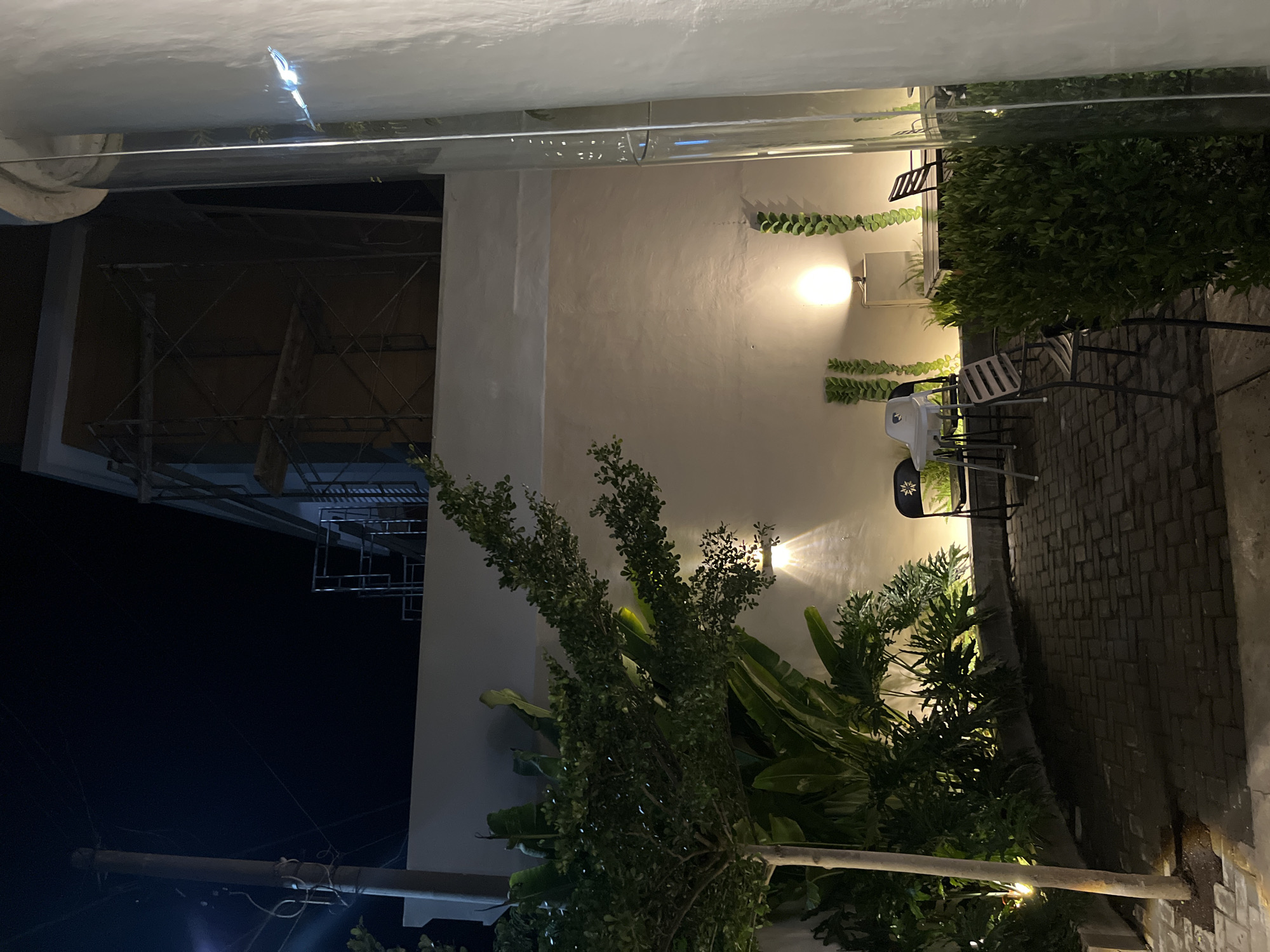 The image depicts an outdoor seating area at night, possibly a cafe or restaurant, in Lhokseumawe, Indonesia. A paved brick patio is visible, furnished with several chairs and a table. One chair is black with a star-shaped cutout, another is a high chair, and a third has a slatted back. Plants and trees, including a small tree with a light-colored trunk, are interspersed throughout the space, some illuminated by small spotlights. Behind the seating area, a light-colored wall is visible, with a large globe-shaped light fixture casting a warm glow. Further back, scaffolding suggests ongoing construction on a building. The scene is enclosed by a wall and a tall, clear glass structure on the right. The overall ambiance is dimly lit and serene.