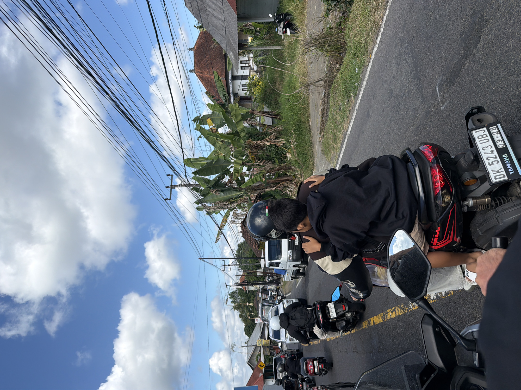 The image depicts a congested roadway in Baturiti, Indonesia, with multiple motorcycles and vehicles on a paved road under a partly cloudy sky. In the foreground, two individuals are riding on a motorcycle, with the rear passenger, a child, embracing the driver. The driver is wearing a black helmet and a black jacket with white and tan accents. The child is wearing a black hoodie. The motorcycle has a black license plate reading "DK 5243 UBJ" and "YAMAHA 03.28". Other motorcycles, cars, and a white truck are visible further down the road, suggesting traffic. The surrounding environment includes a grassy verge, banana trees, and houses with tiled roofs. Electrical wires are strung across the scene.