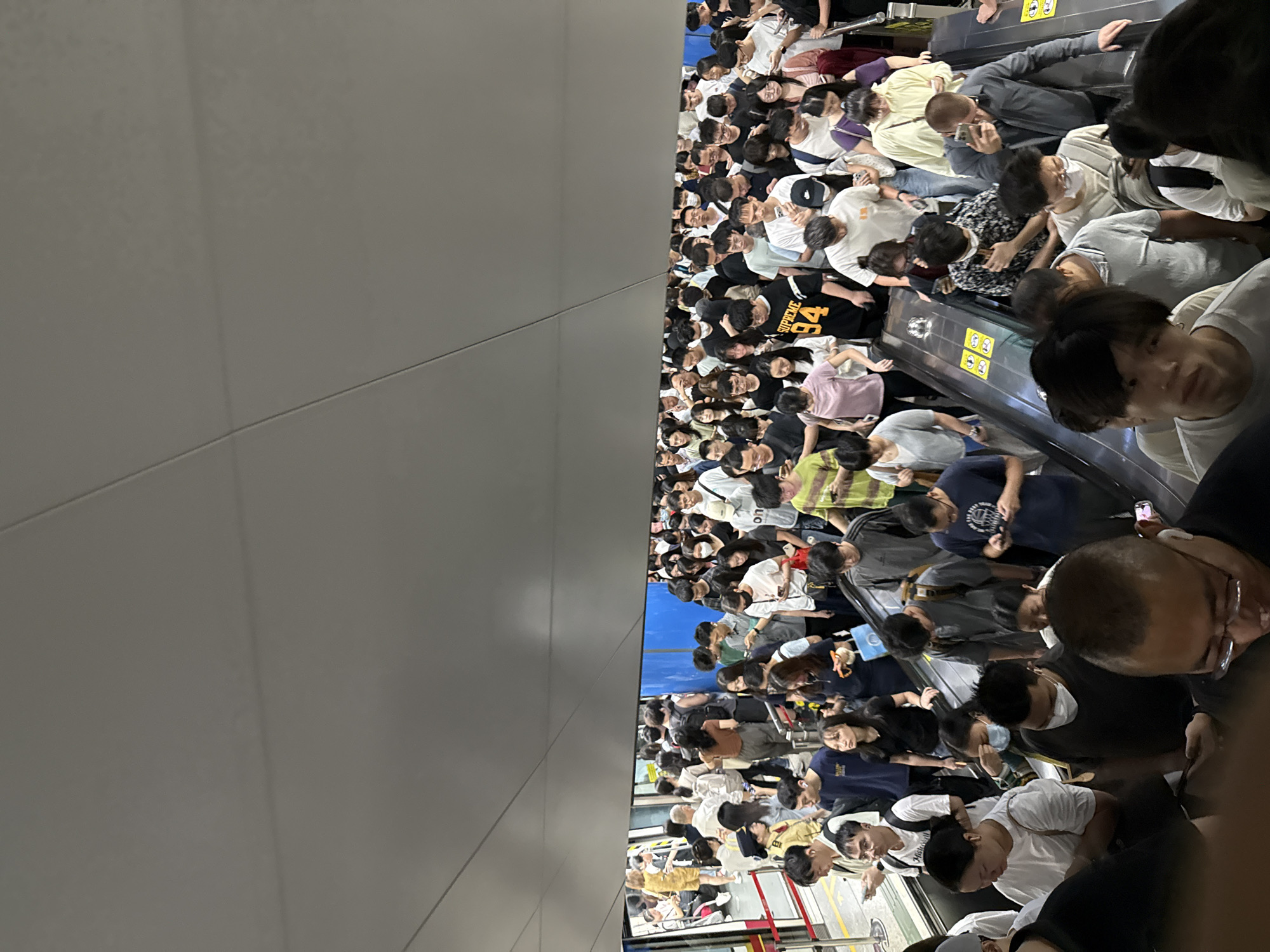 A crowded escalator in Haizhu District, China, shows dozens of people ascending. The environment appears to be a subway station, with polished floors and gray walls and ceiling. People are dressed casually in t-shirts and jeans. Many are looking at their phones, while others engage in conversation or simply wait to reach the top. The overall impression is one of urban transit, with the movement of people and the sense of a busy day.