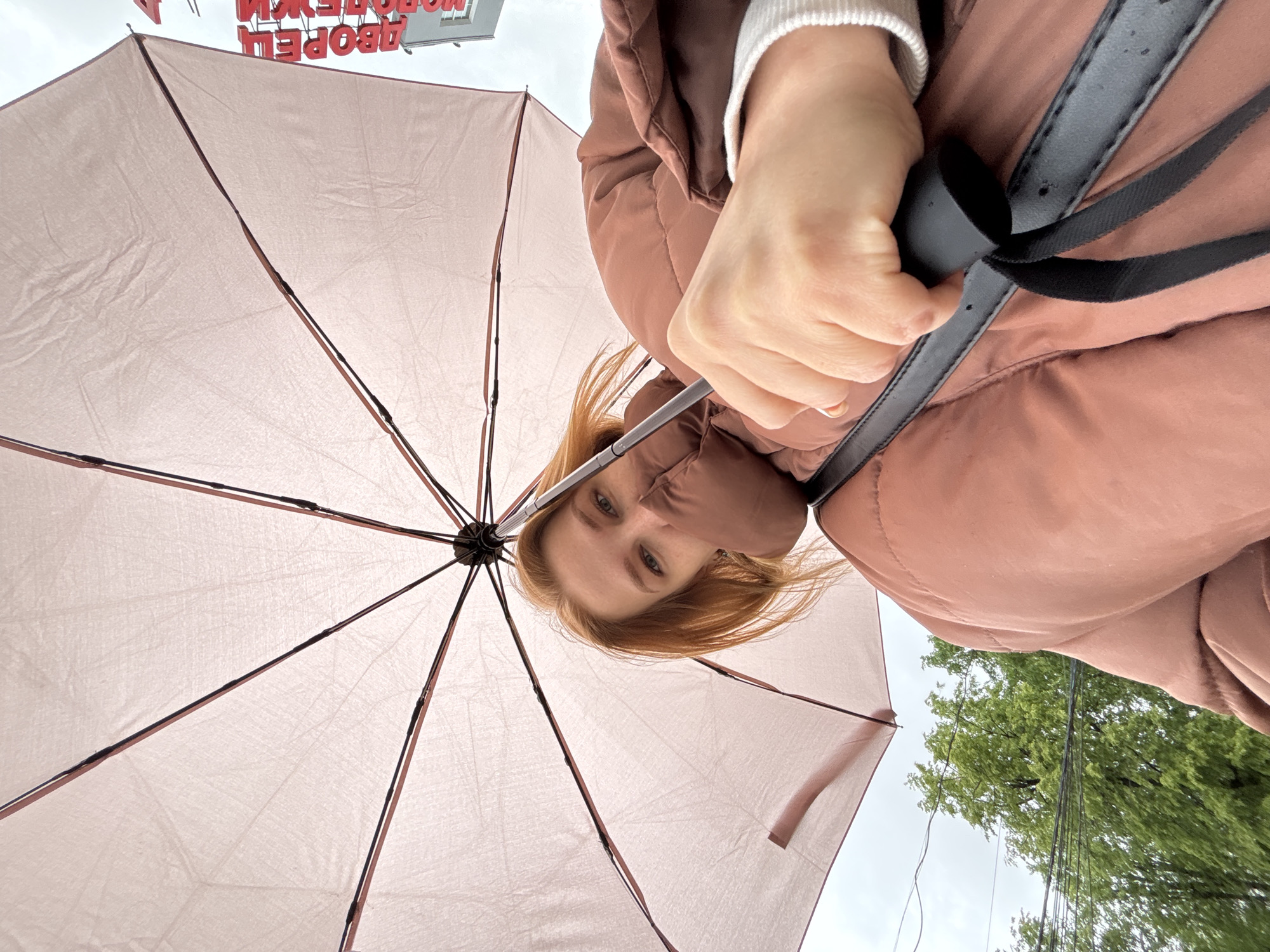 A low-angle shot captures a person holding a pink umbrella. The individual wears a puffy, brown jacket and a white sweater underneath. Their ginger hair is visible beneath the umbrella. The background includes a view of the sky and a building sign with upside-down Russian script that reads "ДВОРЕЦ МОЛОДЕЖИ," indicating the location is near the Youth Palace in Yaroslavl, Russia. Trees with green leaves are seen in the distance.