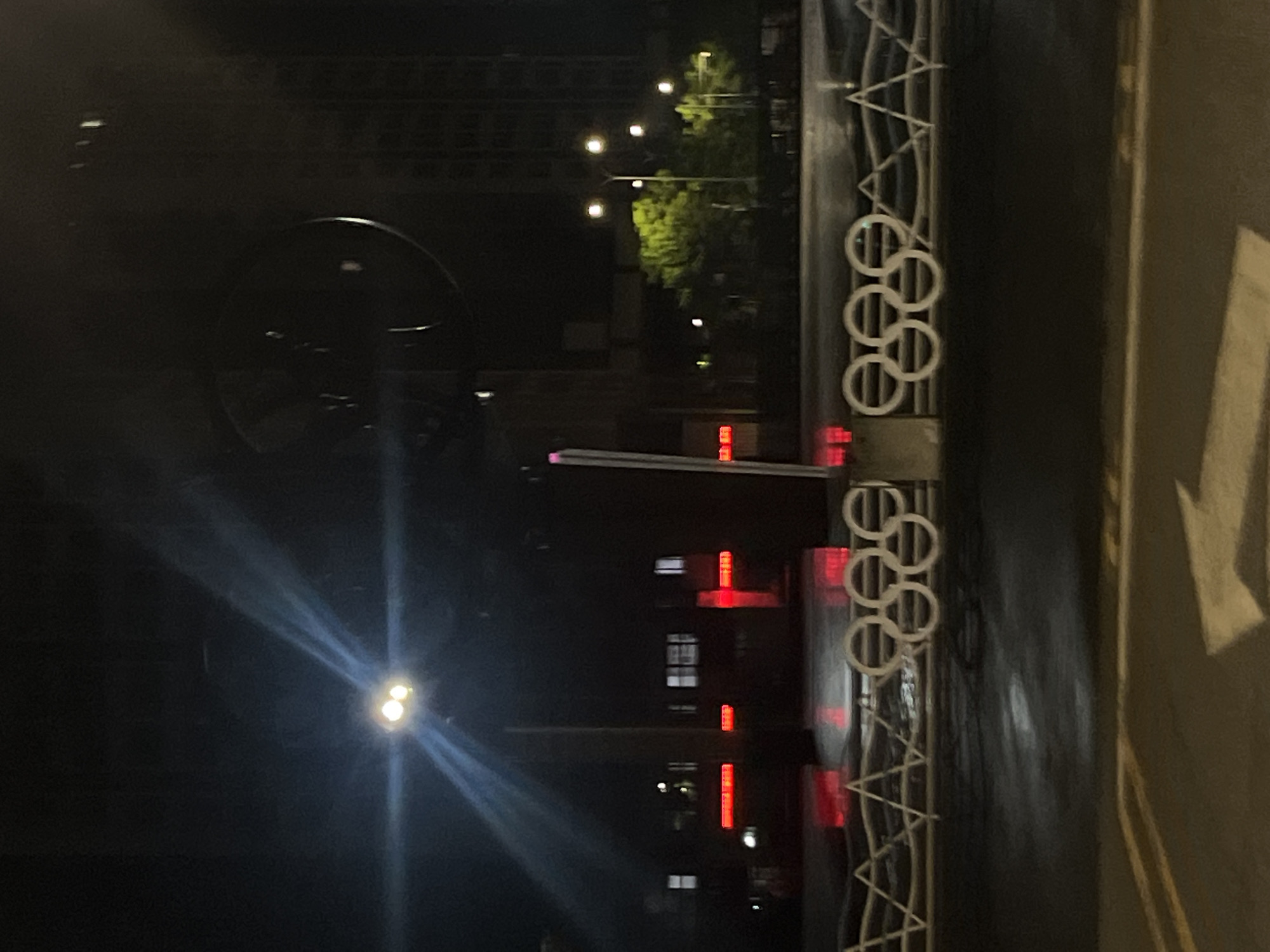 The image depicts an urban street scene at night, likely in Jiulongpo District, China, characterized by artificial lighting and wet pavement. In the foreground, a dark asphalt road features a prominent white arrow painted on its surface, indicating a forward turn. Above the road, a white decorative fence or barrier spans the width of the frame, prominently displaying the five interconnected Olympic rings. To the left of the Olympic rings, the barrier features a pattern of interconnected triangles.
In the middle ground, multiple bright streetlights emit strong beams and lens flares, illuminating the immediate area. Below these lights, several rectangular red illuminated signs or light fixtures are visible along what appears to be a median or structure. Further in the background, additional streetlights cast a green glow on several trees. Beyond the trees and structures, dark, multi-story buildings are visible, some with illuminated windows. The wet appearance of the road surface suggests recent rain or damp conditions. No individuals or vehicles are explicitly visible.