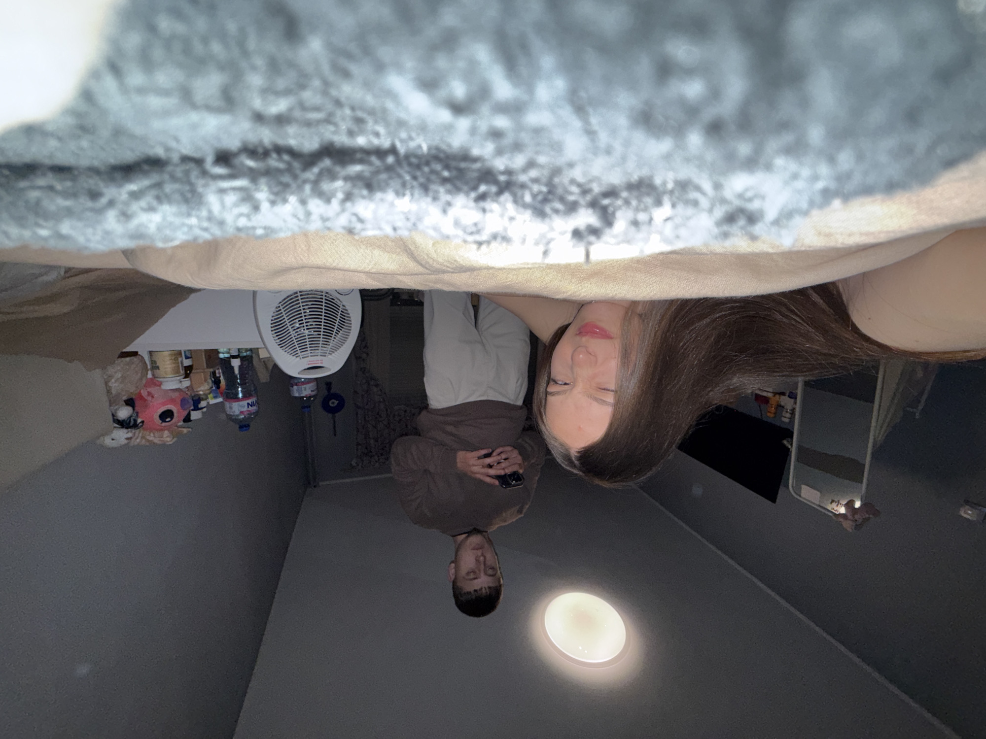 This image, set in Sofia, Bulgaria, captures a low-angle perspective of two individuals and a room's interior. A fluffy grey blanket or textile drapes across the top of the frame. Below it, a person with long, dark brown hair and a relaxed expression is visible on the right, looking upwards. To their left and slightly further back, another person with short dark hair is seated or lying down, wearing a brown long-sleeved top. This individual is holding a dark smartphone with both hands, focusing their attention on it.
The background consists of plain dark grey walls. On the left side of the room, a white oscillating fan is visible on a surface, alongside various items including a pink stuffed animal (resembling Stitch), bottles (one with partially visible text like "NIVA"), and jars. In the lower right corner, a bright, circular ceiling light fixture illuminates the space. A mirror on the right wall reflects a dark segment of the room. The overall scene suggests a casual, indoor setting, possibly an evening gathering, given the reliance on artificial lighting.