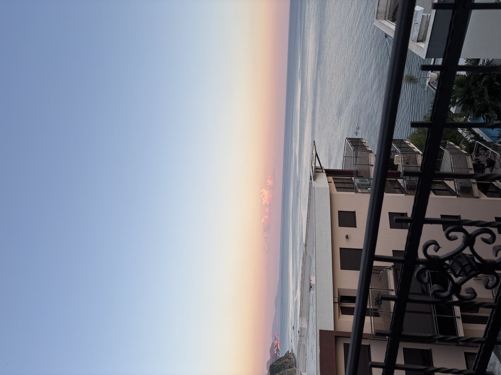 The image depicts a serene coastal scene at dawn or dusk, likely from a balcony in Budva, Montenegro. The sky transitions from a pale blue at the top to soft pinks and oranges near the horizon, where a few scattered clouds are illuminated by the rising or setting sun. The calm sea stretches out to meet the sky, with a distant coastline visible on the left, characterized by gentle hills and some illuminated structures.
In the foreground, a light pink, multi-story building with balconies and dark-shuttered windows dominates the lower portion of the frame. A black metal railing with ornate detailing is visible across the bottom, suggesting the viewpoint is from a balcony or terrace. Several small boats are scattered on the water. Below the main building, glimpses of a swimming pool and palm trees can be seen. The overall atmosphere is tranquil and picturesque.