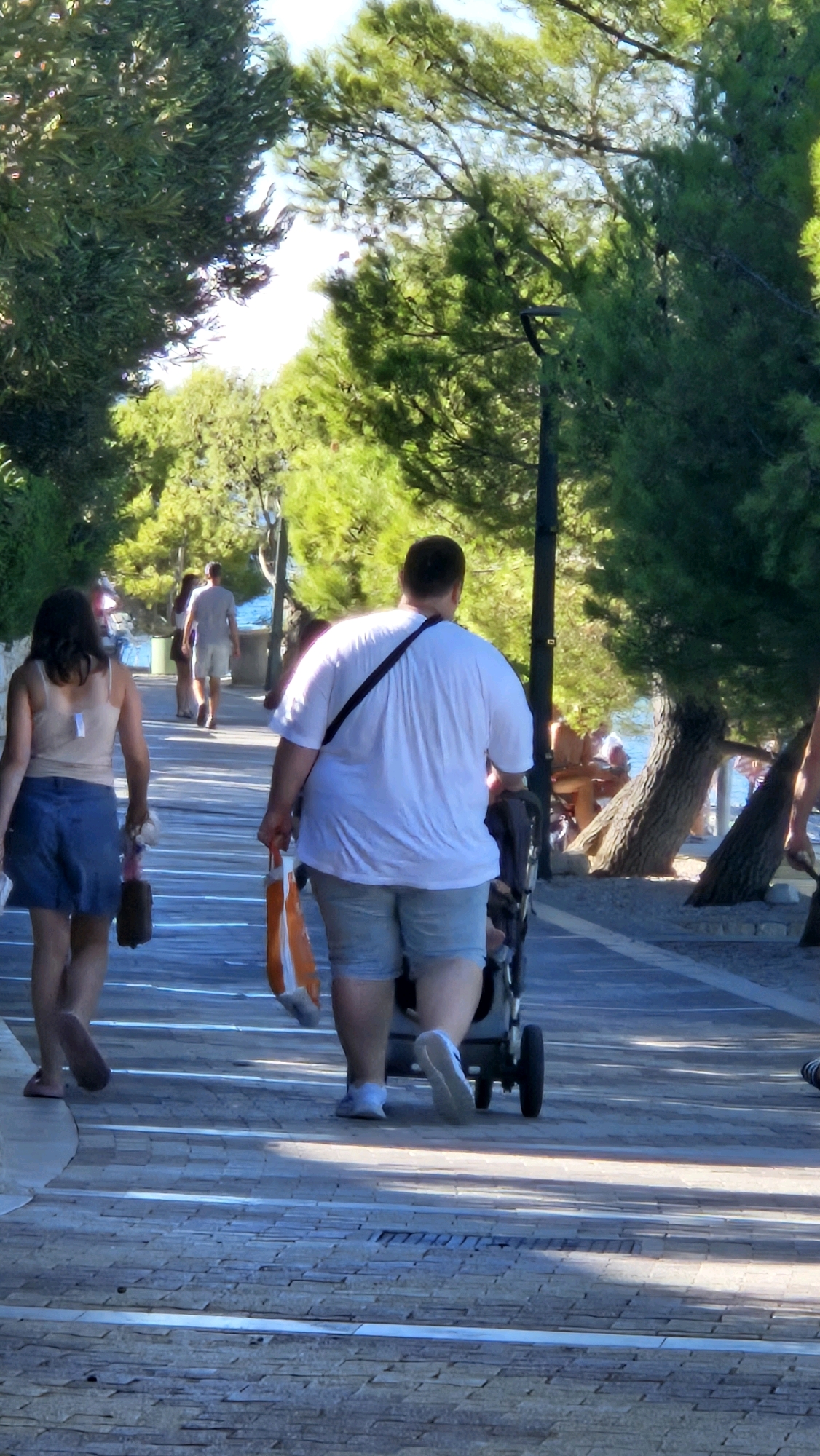 The image depicts a paved walkway lined with trees, likely in a coastal town or resort area. In the foreground, a woman with dark hair is walking away from the camera, wearing a beige tank top and a blue denim skirt. She is carrying a dark brown bag in her right hand. Beside her, walking in the same direction, is a man with short brown hair, wearing a white t-shirt and light blue shorts. He is pushing a dark-colored stroller with wheels and carrying an orange and white bag over his left shoulder. In the background, other individuals are visible, including a man in a gray t-shirt and shorts walking further down the path. The walkway appears to be made of stone or concrete pavers, and dappled sunlight filters through the trees overhead. The overall setting suggests a warm, sunny day. The presence of the walkway, trees, and proximity to what appears to be water (in the background) is consistent with the description of Općina Tučepi, Croatia, a coastal municipality known for its beaches and scenic promenade.