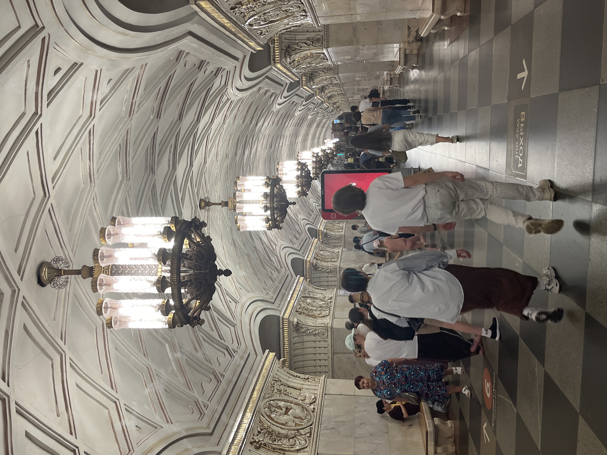 The image depicts the interior of a Moscow Metro station, likely in Russia. The station features a grand architectural style with a high, vaulted ceiling adorned with intricate geometric patterns and decorative moldings. Several large, ornate chandeliers hang from the ceiling, casting a bright, warm light. The walls are lined with marble and decorated with bas-relief sculptures within arched alcoves, gilded trim, and ornate columns.
The floor is laid with a checkerboard pattern of dark and light tiles. People are seen walking along the platform and sitting on benches. The individuals are dressed in casual summer attire, suggesting warm weather. Some people are walking towards the camera, while others are walking away. One person in the foreground is wearing a light-colored striped outfit and is walking away from the camera. Another person in the mid-ground is wearing a light blue striped shirt and a dark skirt, also walking away. In the background, more people are visible, some in small groups, suggesting a moderately busy public space.
A red digital screen is visible on a column in the middle distance. Signs with arrows and text in Russian, including what appears to be "ВЫХОД" (Exit) and "WAY OUT", are visible on the floor, indicating direction. The overall impression is one of opulence and grandeur characteristic of historic Moscow Metro stations.
