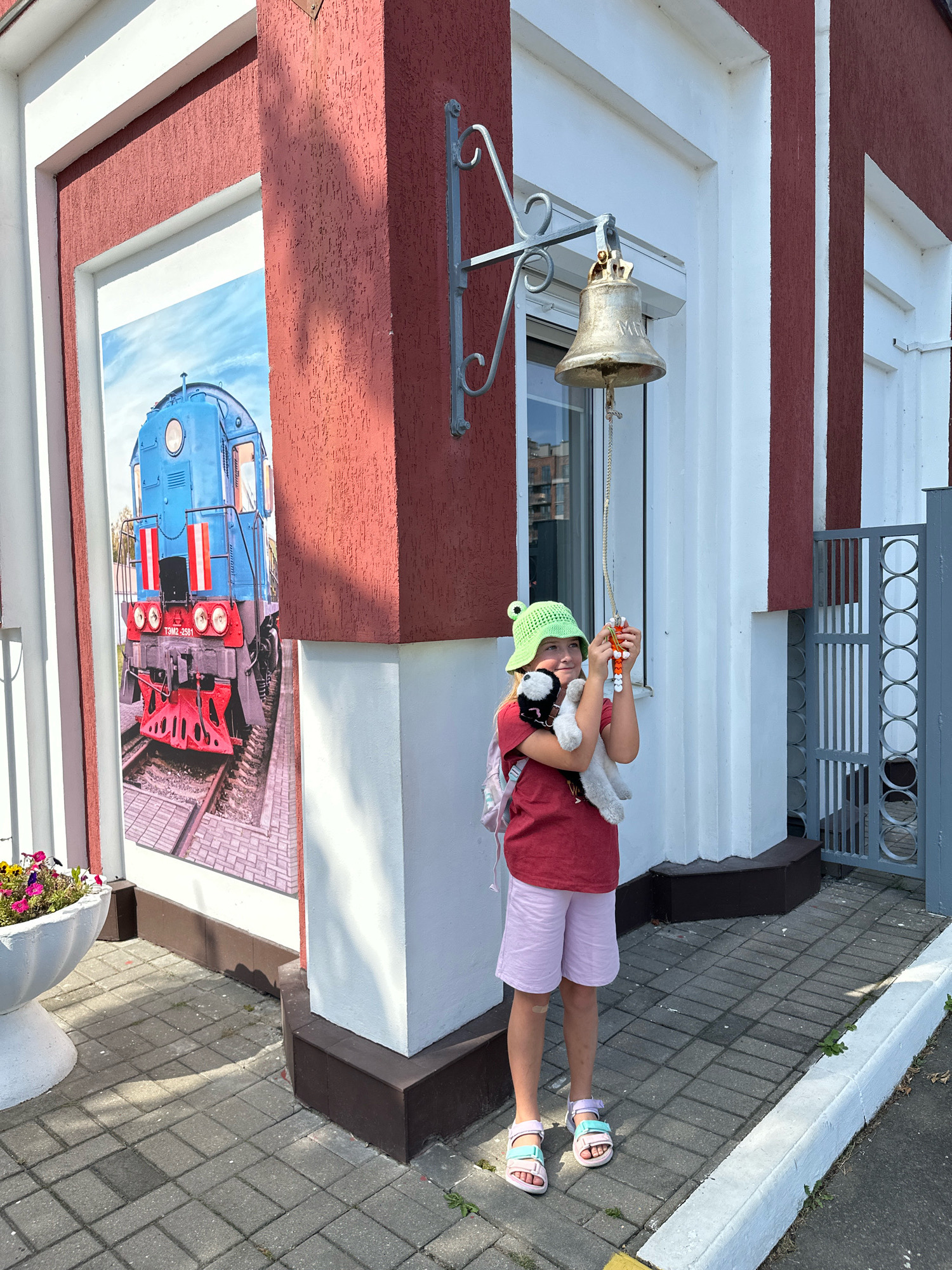 The image depicts a young girl holding a stuffed animal and a small bell with orange beads, standing in front of a building with a mural of a blue train. She is wearing a green bucket hat, a red t-shirt, pink shorts, and sandals. The building has white and dark red walls, and a metal bell is hanging from a decorative bracket on the wall. The ground is paved with grey interlocking bricks. A white planter with flowers is visible to the left of the building. The scene appears to be outdoors on a sunny day. This location is likely a museum or attraction related to trains in Brest, Belarus.