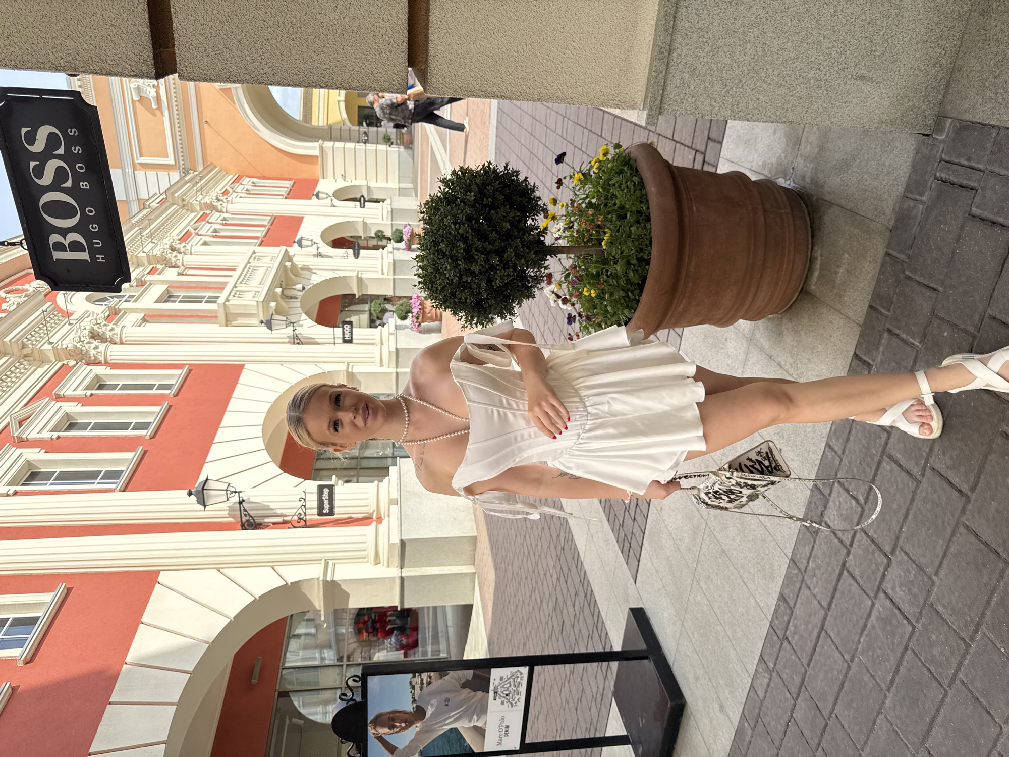 The image depicts a young woman posing outdoors, likely in a shopping district given the presence of storefronts and signage. She is wearing a white dress and white sandals, and is holding a patterned clutch bag. A large terra cotta pot with a small tree and flowers sits behind her. In the background, a building with red and white architectural details is visible, along with a "BOSS Hugo Boss" sign. The scene appears to be during the daytime, with bright lighting suggesting fair weather. The woman's expression is a slight smile, conveying a relaxed and confident demeanor. The location is identified as Saint Petersburg, Russia.