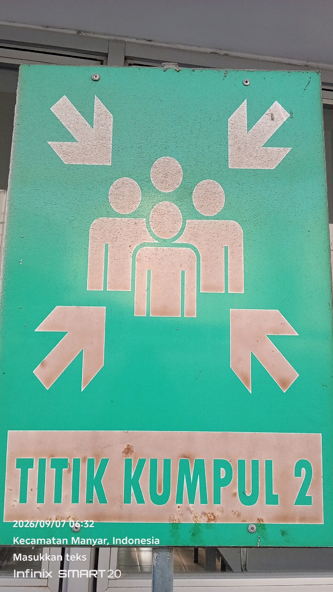 A green sign with white graphics indicates a meeting point. Three stylized figures, representing people, are depicted in the center. Four white arrows point towards the figures, two from the top and two from the bottom. Below the graphics, a sign reads "TITIK KUMPUL 2" (Assembly Point 2) in white text on a brown background. Further down, text indicates the date and time "2026/09/07 06:32", the location "Kecamatan Manyar, Indonesia", and "Infinix SMART 20". The sign appears to be mounted outdoors, with a gray metal pole visible in the lower right corner. The background suggests an industrial or institutional setting with a concrete wall and ceiling.