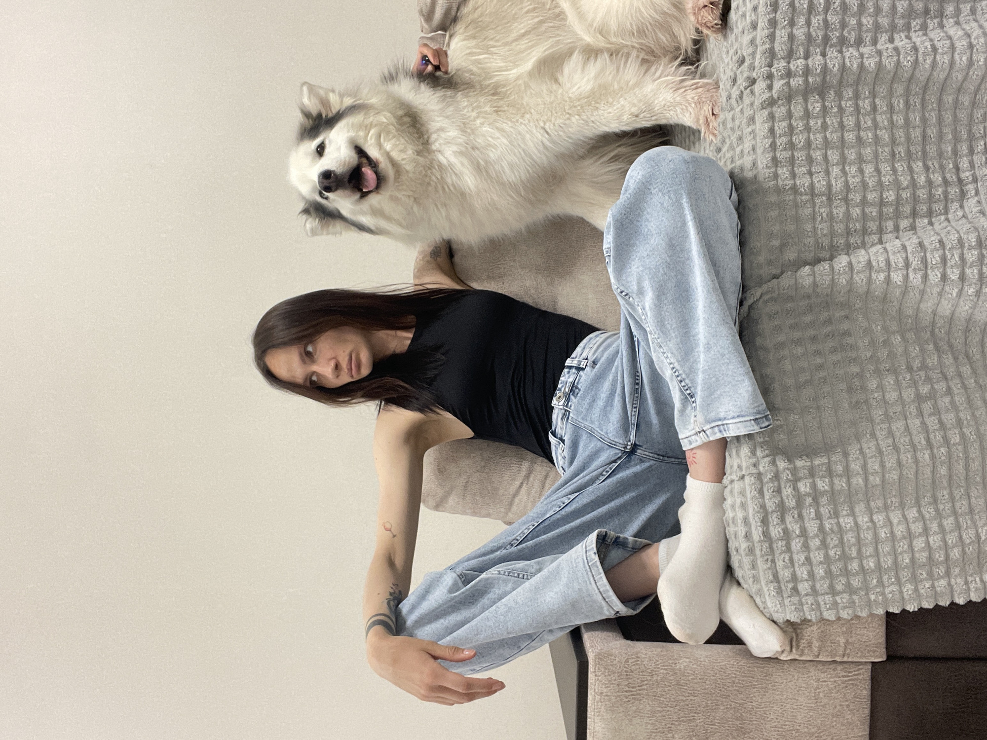 A young woman with dark hair and fair skin is lying on a couch with a large, fluffy dog. The woman is wearing a black tank top and light blue jeans. She has tattoos on her arms. The dog is white and gray with a pink tongue sticking out. The couch is covered with a gray, textured blanket. The background is a plain, white wall. The scene appears to be indoors, possibly in a home. There is no information available in the image to indicate the location is Pervomaysky, Russia.