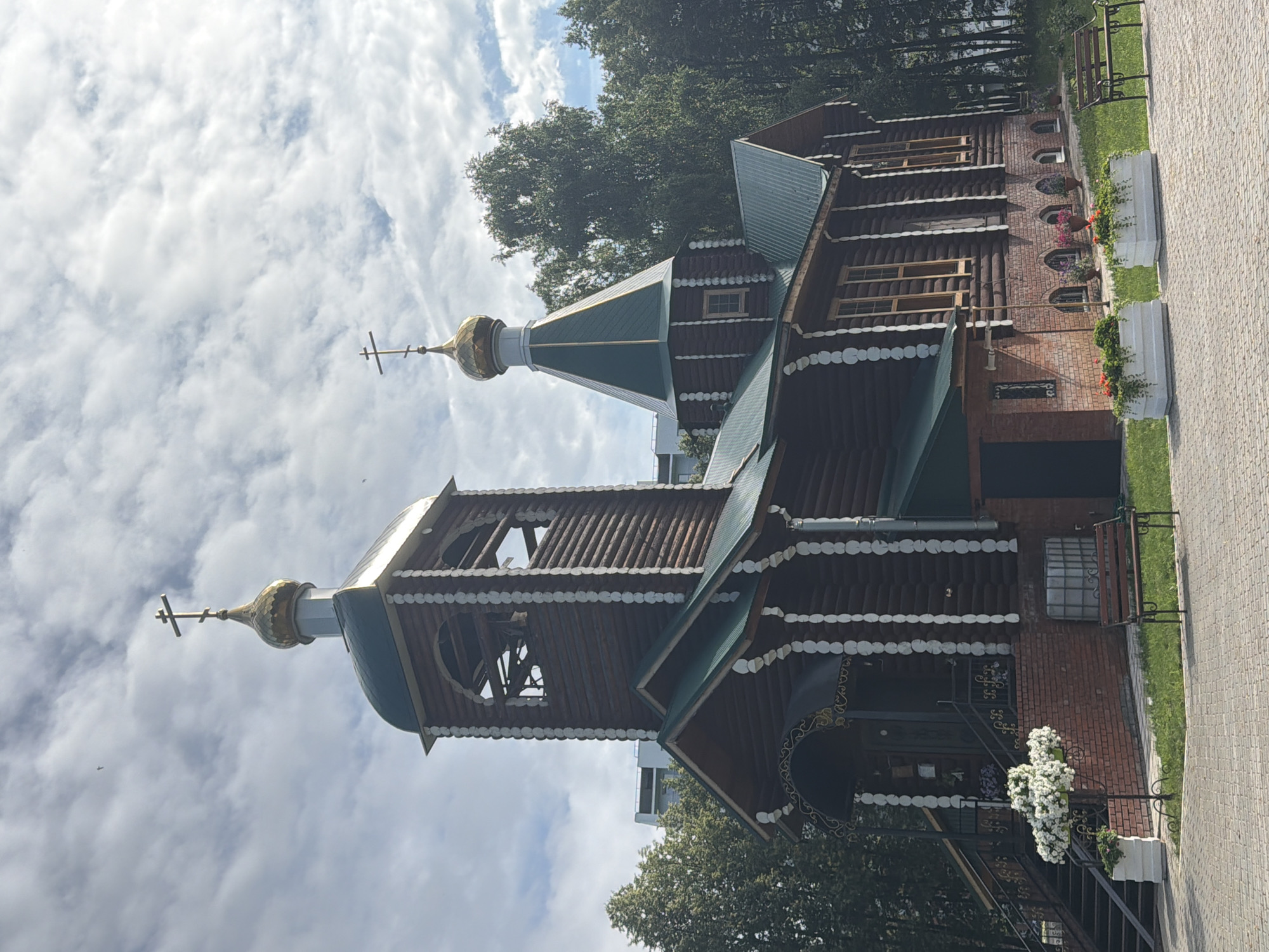 The image shows a wooden Orthodox church with a bell tower and a central dome, located in Chaikovsky, Russia. The church is constructed from dark logs with white decorative trim. The bell tower has arched openings, and the roof is green. A smaller green-roofed section with a golden dome is visible to the right of the bell tower. The church is set against a backdrop of green trees and a cloudy sky. In front of the church, there are stairs leading to an entrance, flanked by white planters with white flowers. A brick base supports the wooden structure, and a paved walkway with a bench is visible in the foreground. Several flower planters with red and pink flowers are placed along the grassy area beside the walkway. The scene appears peaceful and well-maintained, suggesting a public or community space.