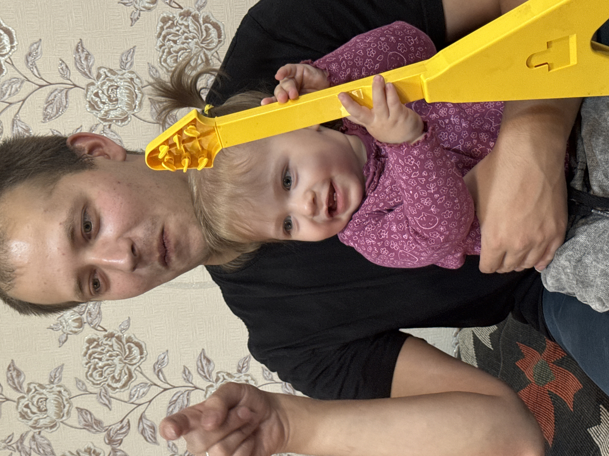 An indoor photograph, likely taken in a home in Nizhnyaya Gar', Russia, captures a man holding a baby. The man, with short dark hair, is seen from the chest up, wearing a black t-shirt. He has a neutral expression and appears to be looking directly at the camera, with his right hand extended and pointing towards the viewer. The baby, nestled in the man's arms, has fair hair, part of which is tied in a small ponytail, and is wearing a purple long-sleeved top with a subtle floral pattern. The baby holds a bright yellow V-shaped toy guitar, looking towards the camera with a wide, happy smile, showing several teeth. The background features light-colored wallpaper adorned with an embossed floral pattern in shades of beige and brown. A patterned blanket or throw with an abstract floral design in red, green, and black is visible on a surface behind the man. The scene conveys a warm, playful family moment. There is no visible text.
