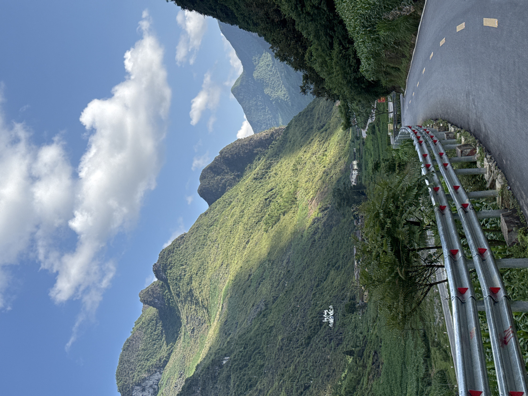 A winding mountain road in Mèo Vạc Commune, Vietnam, is depicted on a sunny day. The road, lined with a metal guardrail featuring red reflectors, curves along a steep, verdant mountain slope. Dense greenery covers the mountain, with a second mountain visible in the background. Patches of sunlight dapple the slope. White clouds dot the blue sky.