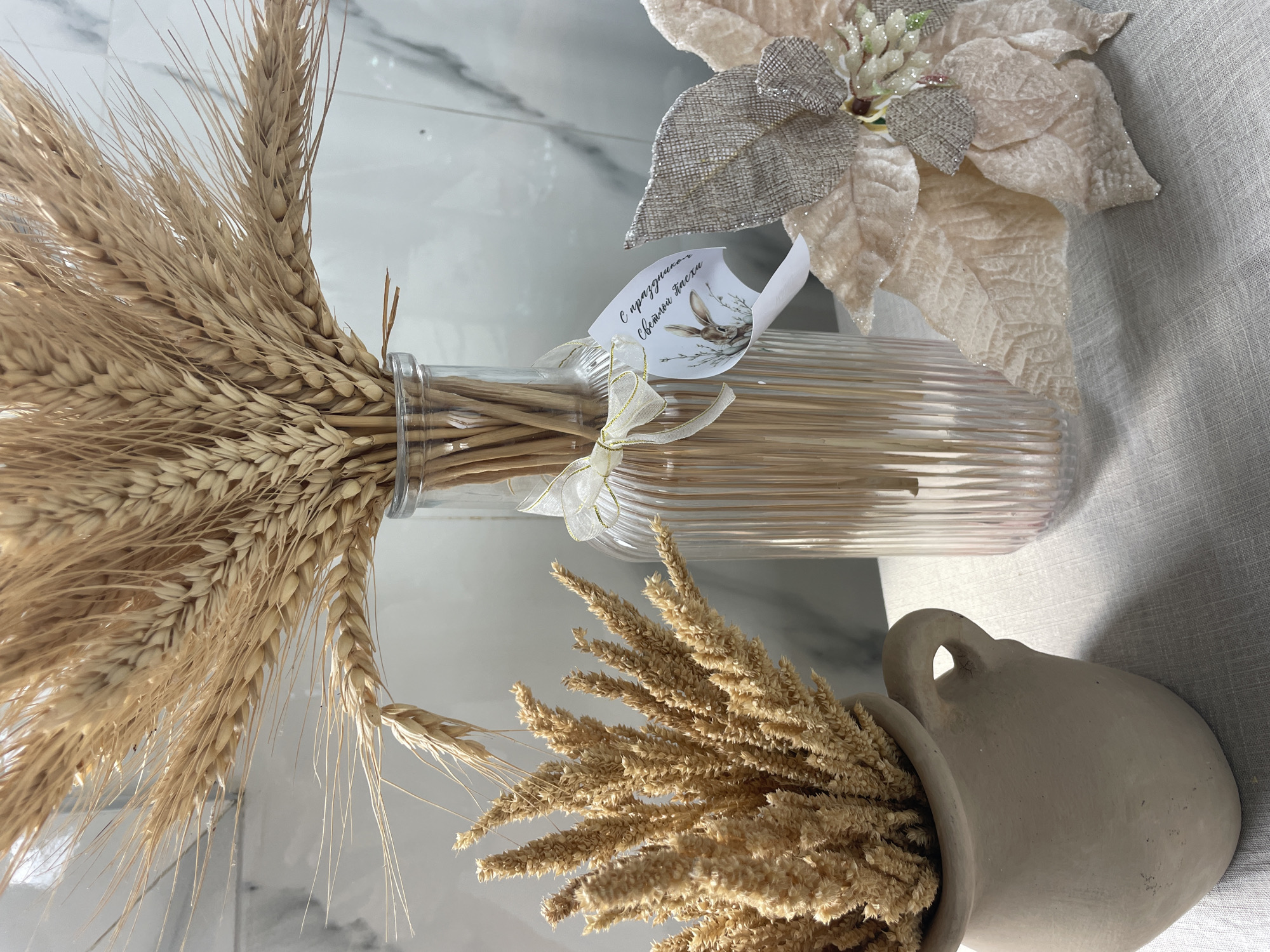 The image shows an arrangement of dried wheat stalks in a clear, fluted glass vase with a white ribbon tied around the neck and a small tag attached. Next to the vase is a clay pot with dried wheat sprigs and a decorative poinsettia flower on a light gray tablecloth. The background features a white marble wall. The location in the city of Unknown, Unknown, cannot be determined from the image.