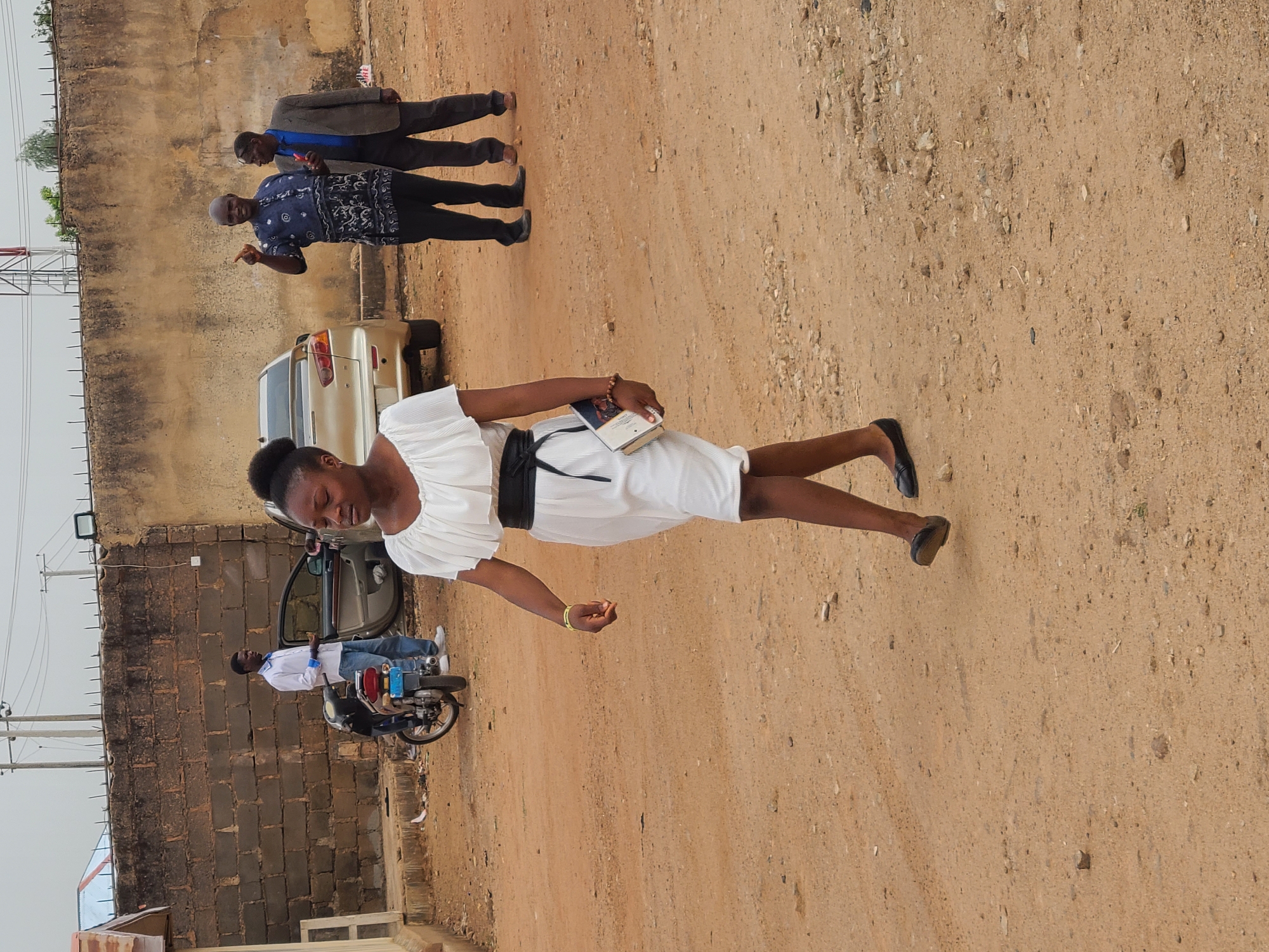 The image depicts an outdoor scene on an unpaved, light brown dirt road in Gura Top, Nigeria, under an overcast daylight sky.
The main subject is a young woman walking forward, dressed in a white short-sleeved dress with a black belt and black flat shoes. She holds a dark-covered book, possibly a religious text, in her left hand, which also features a light-colored bracelet. Her dark hair is styled up.
In the mid-ground, to her left, a man in a white shirt and blue jeans is seated on a dark motorcycle, positioned next to an open car door of a light-colored sedan. Behind him, a tall concrete block wall, topped with barbed wire, runs along the roadside. Power lines and utility poles are visible against the sky.
Further down the road to the woman's right, two other men are standing. One bald man, wearing a blue patterned short-sleeved shirt and dark trousers, is seen gesturing with his right hand. Behind him, another man in a brown suit jacket over a blue shirt and dark trousers stands, appearing to be engaged in conversation or observing the scene.
The ground is composed of compacted dirt mixed with numerous small stones and gravel. The overall scene suggests a casual gathering or movement of people in a semi-urban or residential area. No legible text is clearly visible on any objects within the image.
