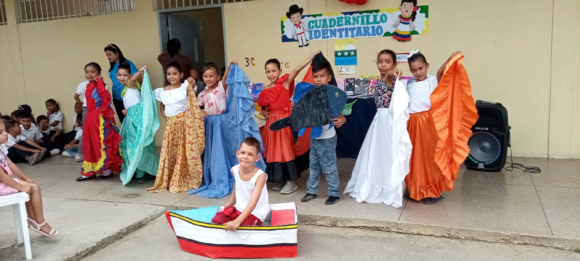 The image shows a group of children in a school or community setting in an unknown city. Ten children are standing in a line, dressed in colorful costumes, possibly for a performance or cultural event. A boy in a white tank top sits in a small, painted boat in front of the line. Another group of children sits on the ground to the left, watching.
Behind the standing children is a pale yellow wall with a doorway and an informational sign above them. An AIWA speaker is to the right of the group. The environment appears to be an outdoor concrete area, likely a courtyard or stage. The photograph has decent lighting, but some details are slightly blurred, indicating a medium-quality camera or a distant shot.