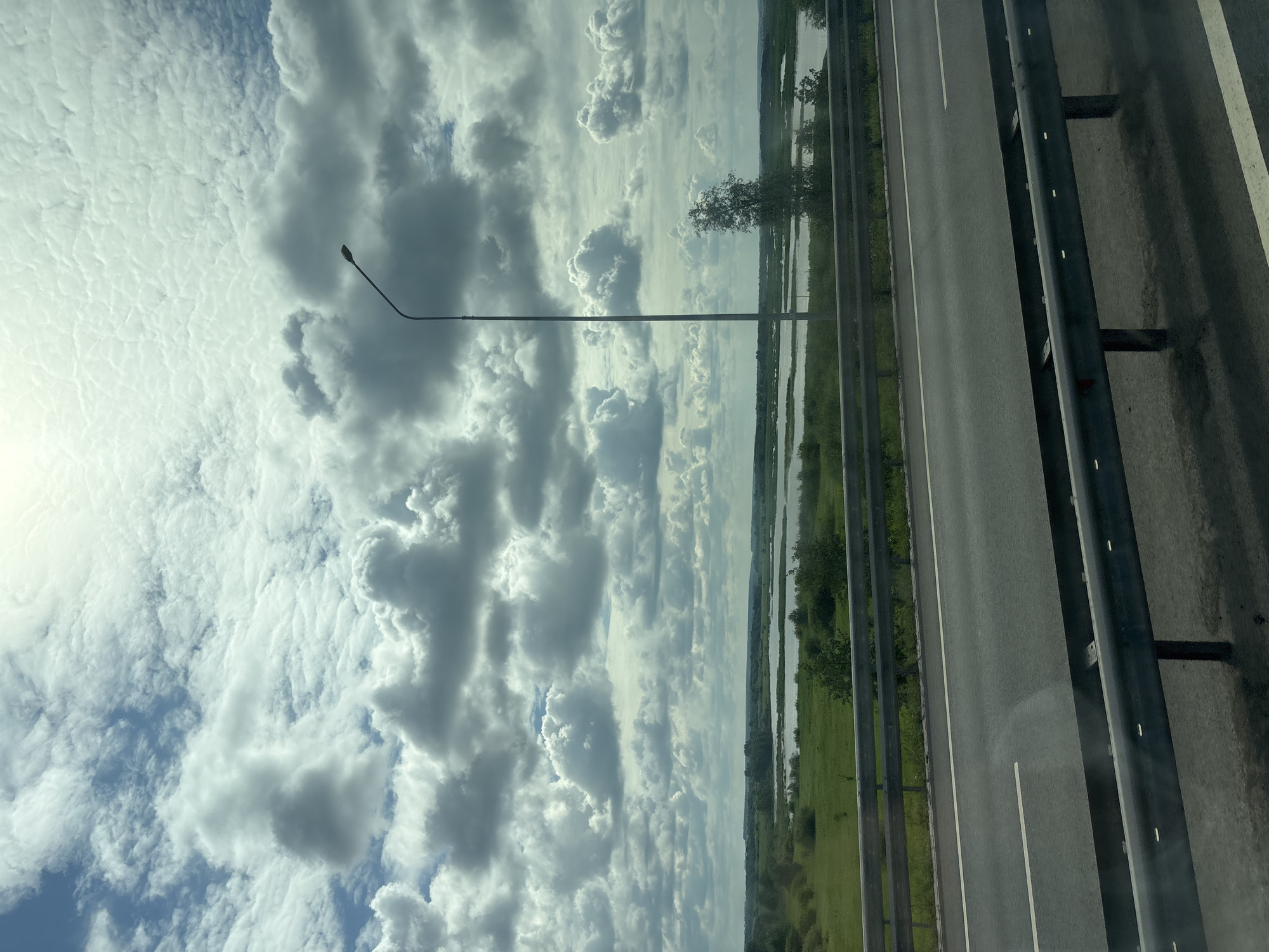 The image captures a view from a highway looking towards a vast landscape under a dramatic, cloud-filled sky. The highway itself is visible in the foreground, featuring asphalt with white lane markings and a metal guardrail. Beyond the highway lies a green grassy field dotted with scattered trees and bushes. Further in the distance, a body of water, likely a river or lake, stretches across the horizon, reflecting the sky. The sky is dominated by a complex pattern of cumulus clouds, with patches of blue sky peeking through. A tall, slender lamppost stands on the right side of the highway, its light fixture pointing upwards. The scene is devoid of any people or discernible activity, presenting a serene, natural environment. It is difficult to definitively place this specific location within Menzelinsk, Russia, without further context or identifiable landmarks.