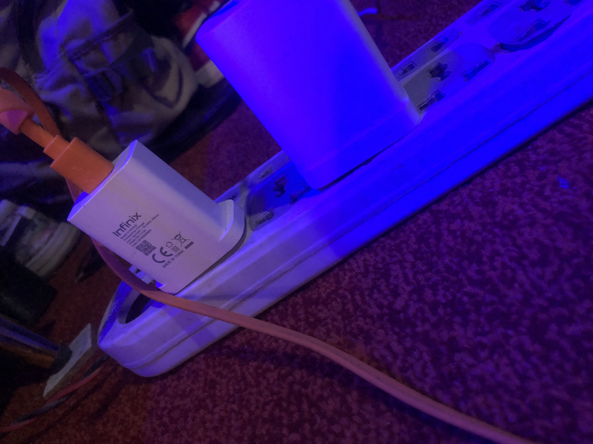 The image depicts a close-up, low-light indoor scene with a strong blue ambient illumination, presumably originating from a plugged-in device. The central subject is an off-white multi-outlet power strip or extension cord resting on a deep red, textured carpet.
An "Infinix" brand white charger, with an orange USB port, is plugged into the leftmost visible outlet of the power strip. Text visible on the charger identifies it as "Model: U100XKA," "Input: 100-240V~ 50/60Hz 400mA," and "Output: 5V==2A," with "MADE IN CHINA" and a "CE" mark. An orange, flat cable extends from the lower-left corner of the frame, running alongside the power strip, and is connected to the orange USB port of the Infinix charger.
Further to the right on the power strip, a cylindrical or rectangular object, possibly a night light or similar electronic device, is plugged in and emits a strong, uniform blue light, dominating the scene's color palette. The outlets on the power strip show signs of use and possibly some discoloration or residue. The carpet beneath is a deep red but appears purple due to the intense blue light. The background is out of focus, revealing indistinct dark shapes that suggest other objects or furniture in a dimly lit room. No people are present. The location is unknown, Unknown.
