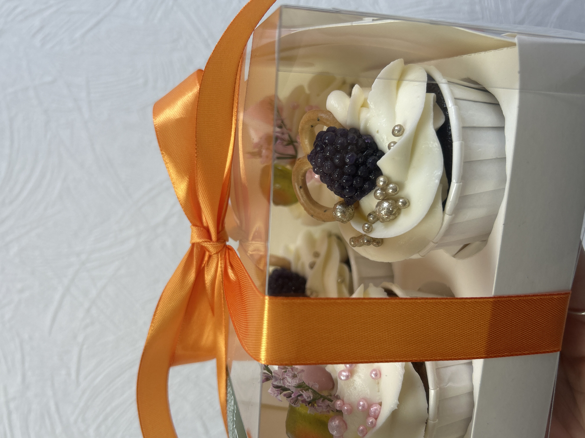 The image shows a clear plastic box filled with cupcakes. Each cupcake has white frosting and is decorated with berries, small pretzels, and silver and pink decorative pearls. An orange ribbon is tied around the box in a bow. The background is a white textured surface. There's no indication of location in Chaikovsky, Russia, or any other location. There are no people visible.