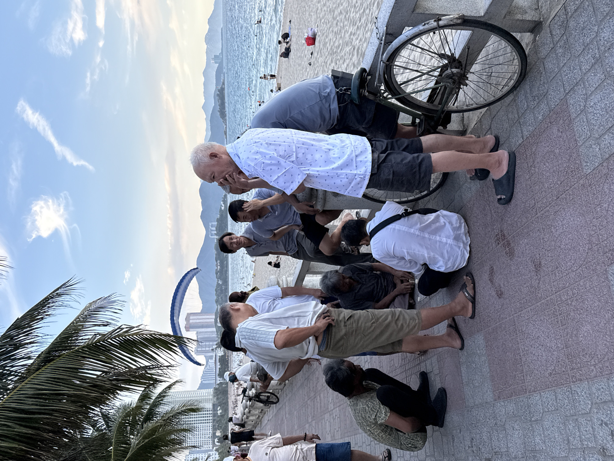 In Nha Trang Ward, Vietnam, a group of men are gathered on a paved promenade overlooking a beach. The beach is populated with people swimming and sunbathing, and a paraglider can be seen in the sky. The men are engaged in various activities; some are conversing, while others are seated on the ground playing a game, possibly chess or checkers. One man stands near a vintage bicycle. The setting appears to be late afternoon or early evening, with the sun casting a warm glow. The promenade is bordered by a low stone wall, and in the background, tall buildings and mountains frame the coastal scene.