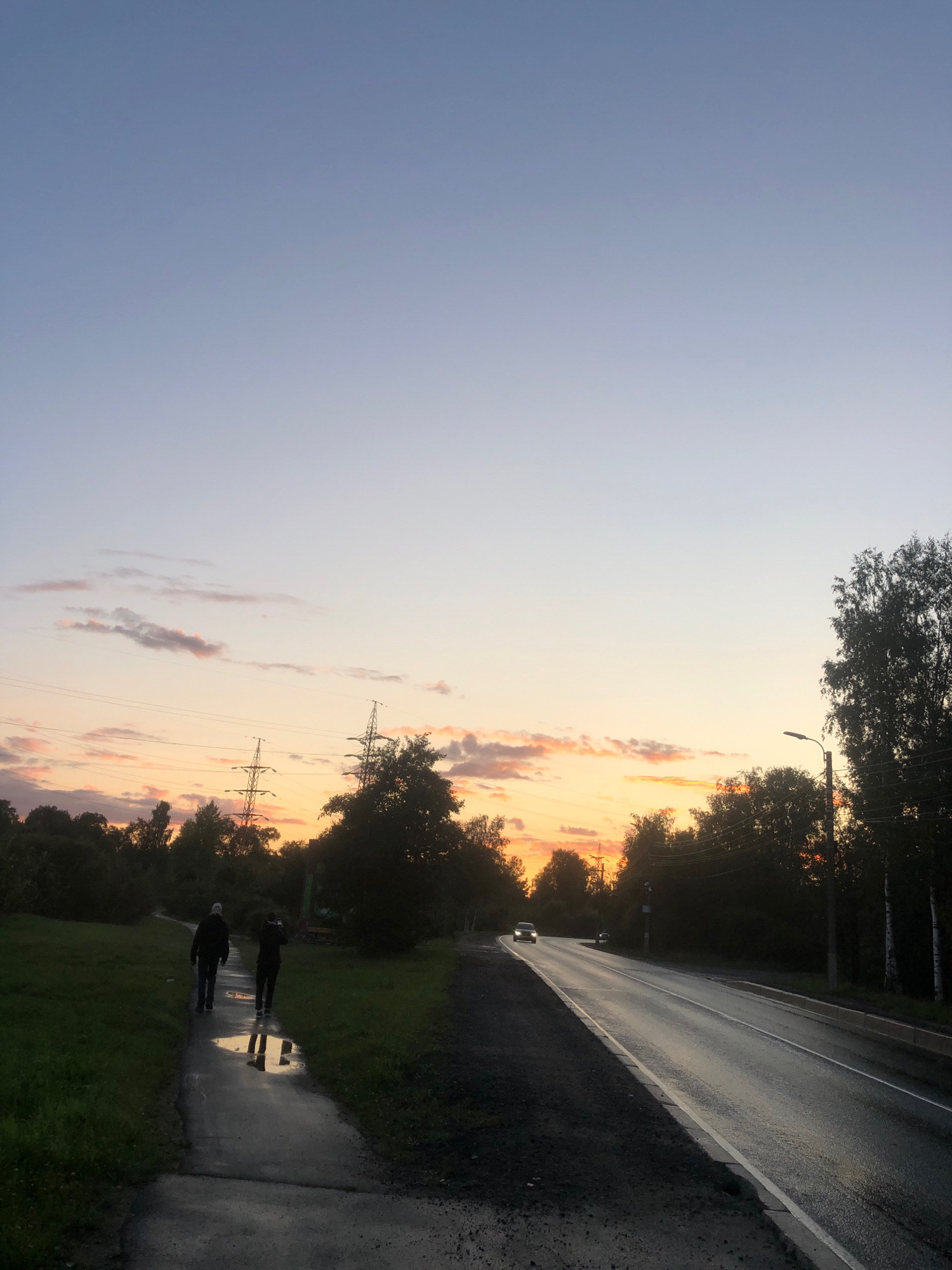 In Beloostrov, Russia, an outdoor scene depicts a road and a walking path at dusk. The sky displays a gradient of pale blue to orange, with wispy clouds. Two individuals are walking away from the camera on the wet, paved walking path. In the background, trees and electrical towers are silhouetted against the sky. A car is driving on the asphalt road, which appears wet from recent rain. The scene captures the atmospheric conditions of twilight in a semi-urban environment.