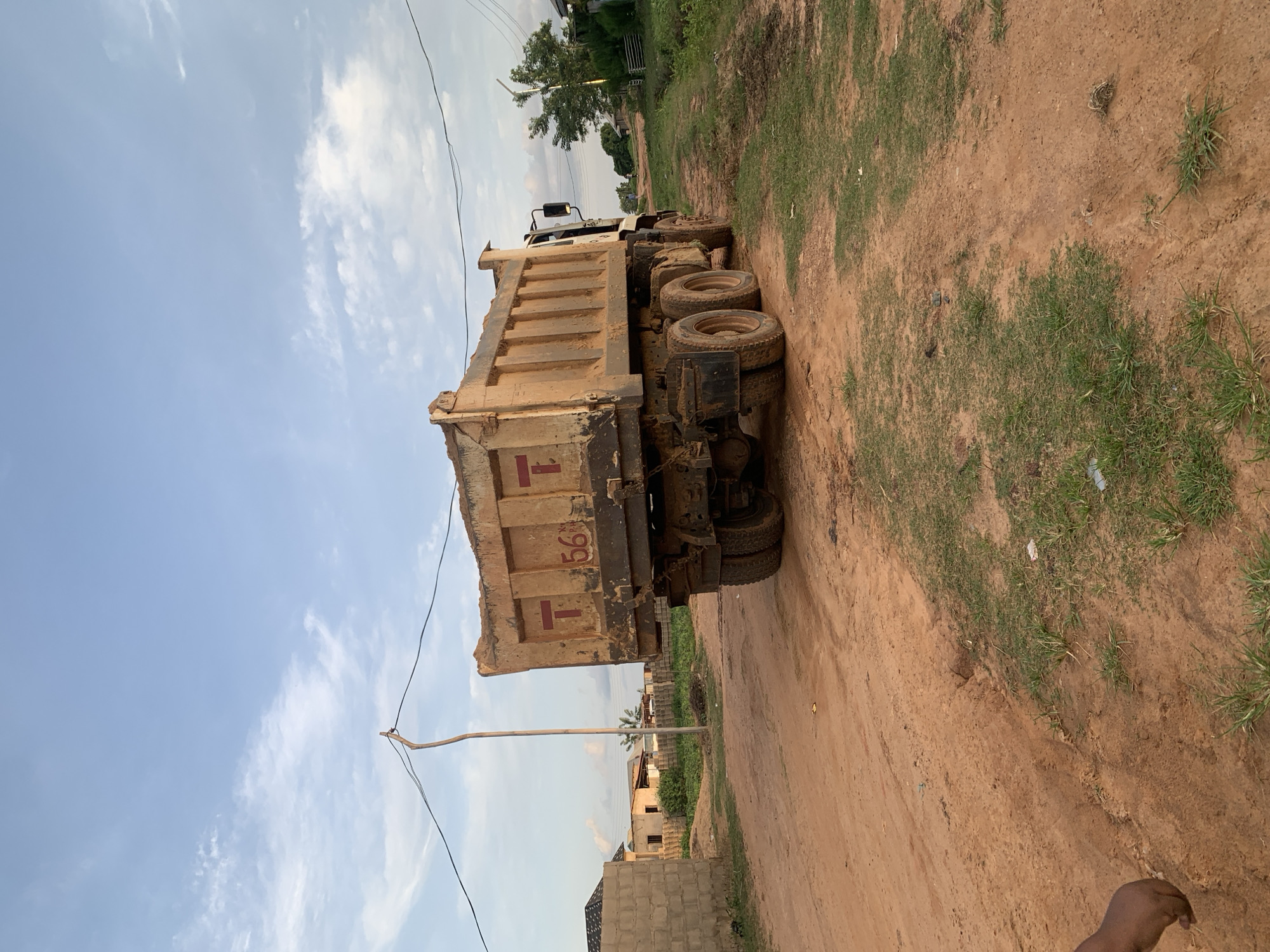 A rear view of a yellow dump truck, laden with sand, is parked on a dirt road in Rido, Nigeria. The truck has the number "56" painted on its side. The road is unpaved and appears to be in a rural or underdeveloped area. To the left of the truck, a partially constructed building with a cinder block wall and a tin roof is visible. In the background, there are other buildings, some with unfinished roofs, and vegetation. A wooden utility pole with power lines runs through the scene. The sky is partly cloudy, with a mix of blue sky and white clouds. The ground is a reddish-brown dirt, with patches of green grass growing in some areas.