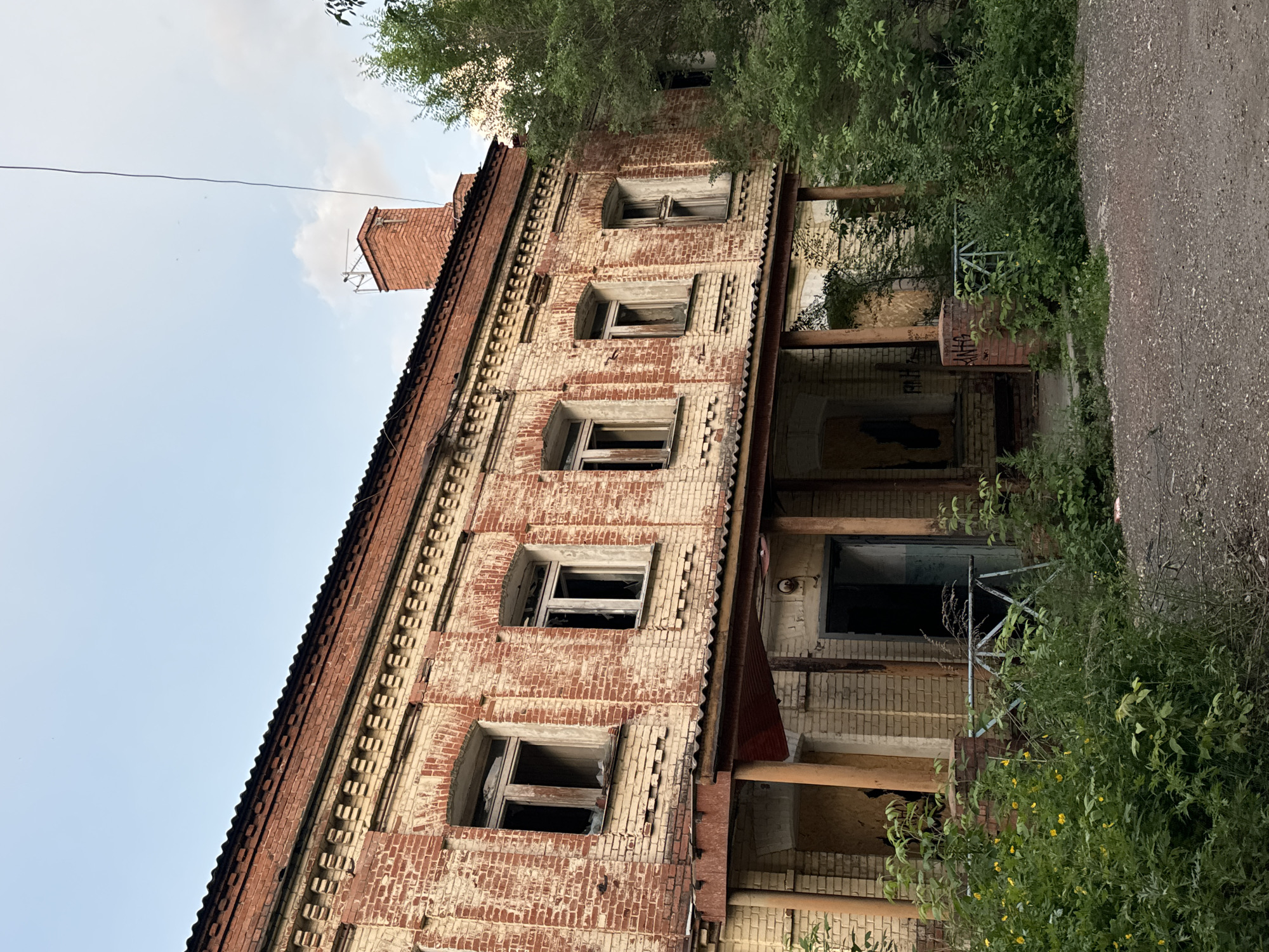 The image depicts the exterior of an abandoned two-story brick building in Krasnoyarsk, Russia, likely a former residential or industrial structure, given its architecture. The building exhibits significant signs of decay. The brickwork is weathered, with portions of the facade appearing discolored or peeling. Several windows on the upper floor are broken or boarded up, and the frames are visibly aged. A corrugated metal roofline is visible, also showing signs of wear.
A covered porch or walkway runs along the ground floor, supported by metal and brick pillars. This area is partially obscured by overgrown vegetation. The entrance to the building appears to be an open doorway, leading into a dark interior. Graffiti is visible on some surfaces, including the boarded-up windows and walls within the covered area.
The surrounding environment is characterized by overgrowth and a general sense of neglect. Lush green foliage, including trees and various plants, encroaches upon the building. Some of these plants are flowering with small yellow blossoms. The ground in front of the building is a rough, gravelly surface, suggesting a neglected pathway or yard. The sky above is partly cloudy, with a pale blue hue. The overall impression is one of an abandoned and dilapidated structure in a state of disrepair, integrated into an overgrown natural setting within a city.